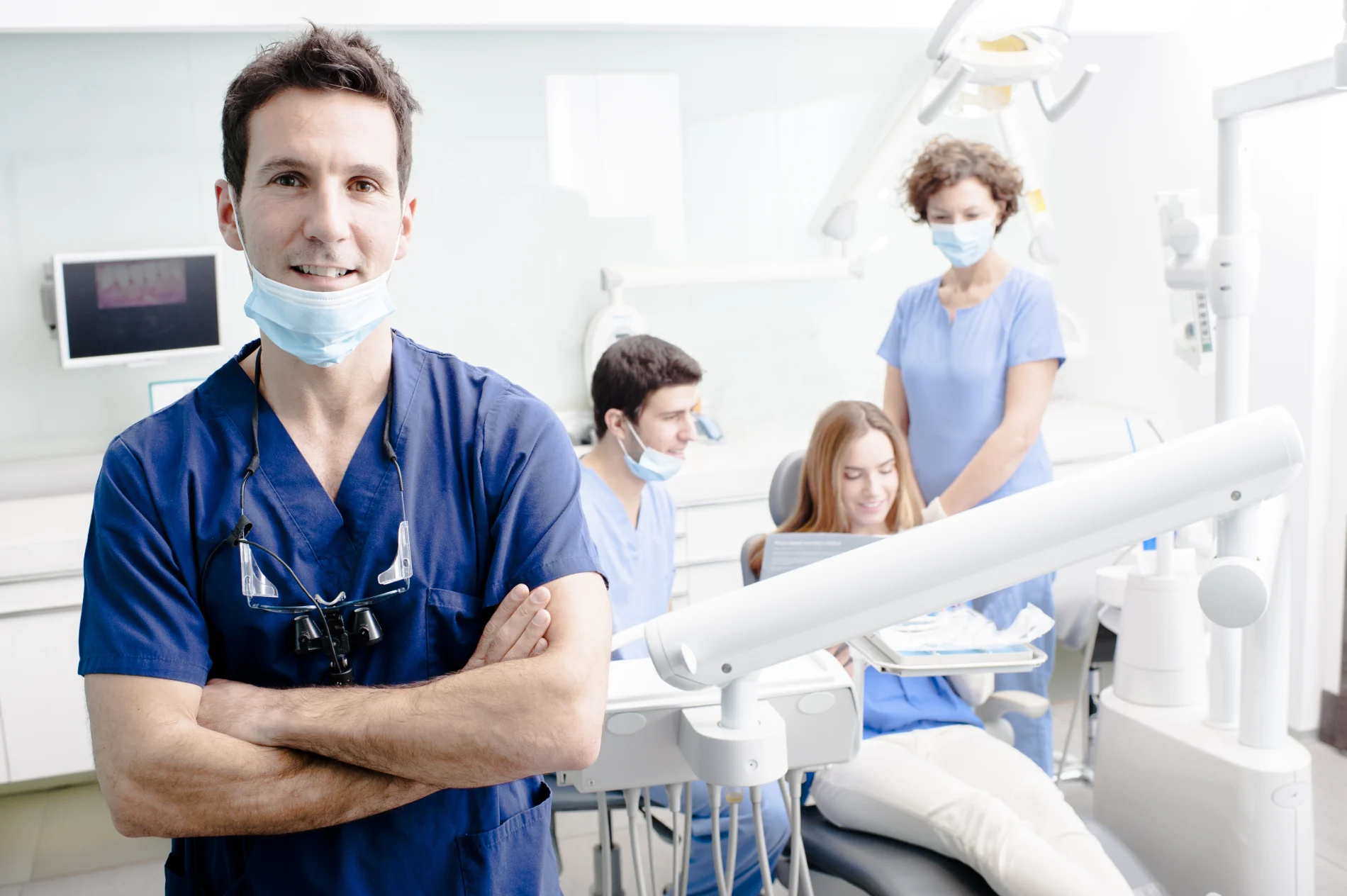 Dentiste de sexe masculin souriant, les bras croisés, avec une équipe de soins travaillant derrière lui.