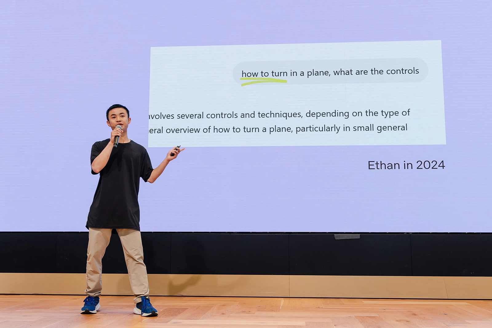 Image of a person speaking into a microphone in front of a large screen displaying a presentation
