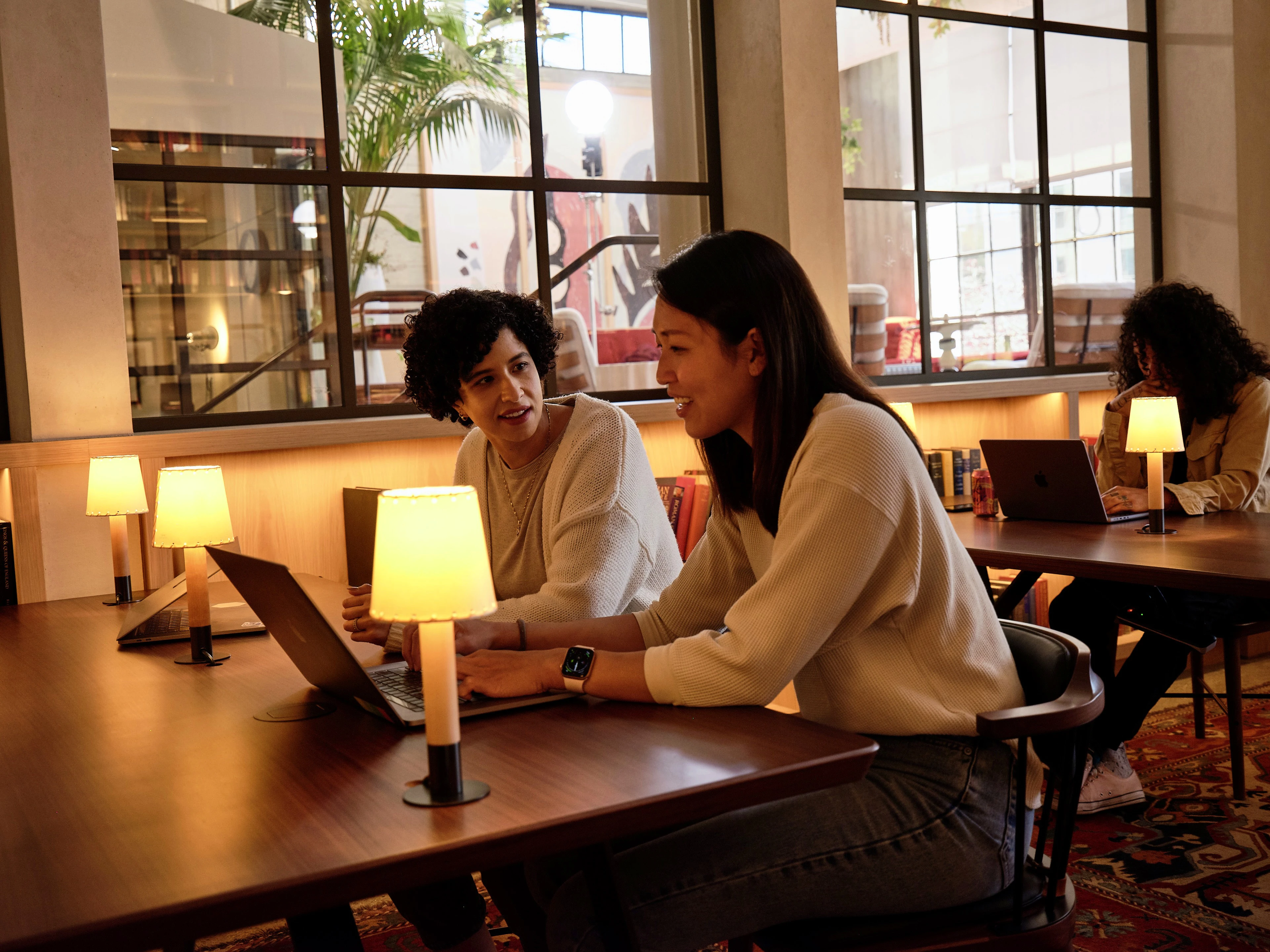 Two women, one with curly hair, are seated at a table working on tablets and conversing in a warmly lit room with table lamps, large windows, and another person in the background.