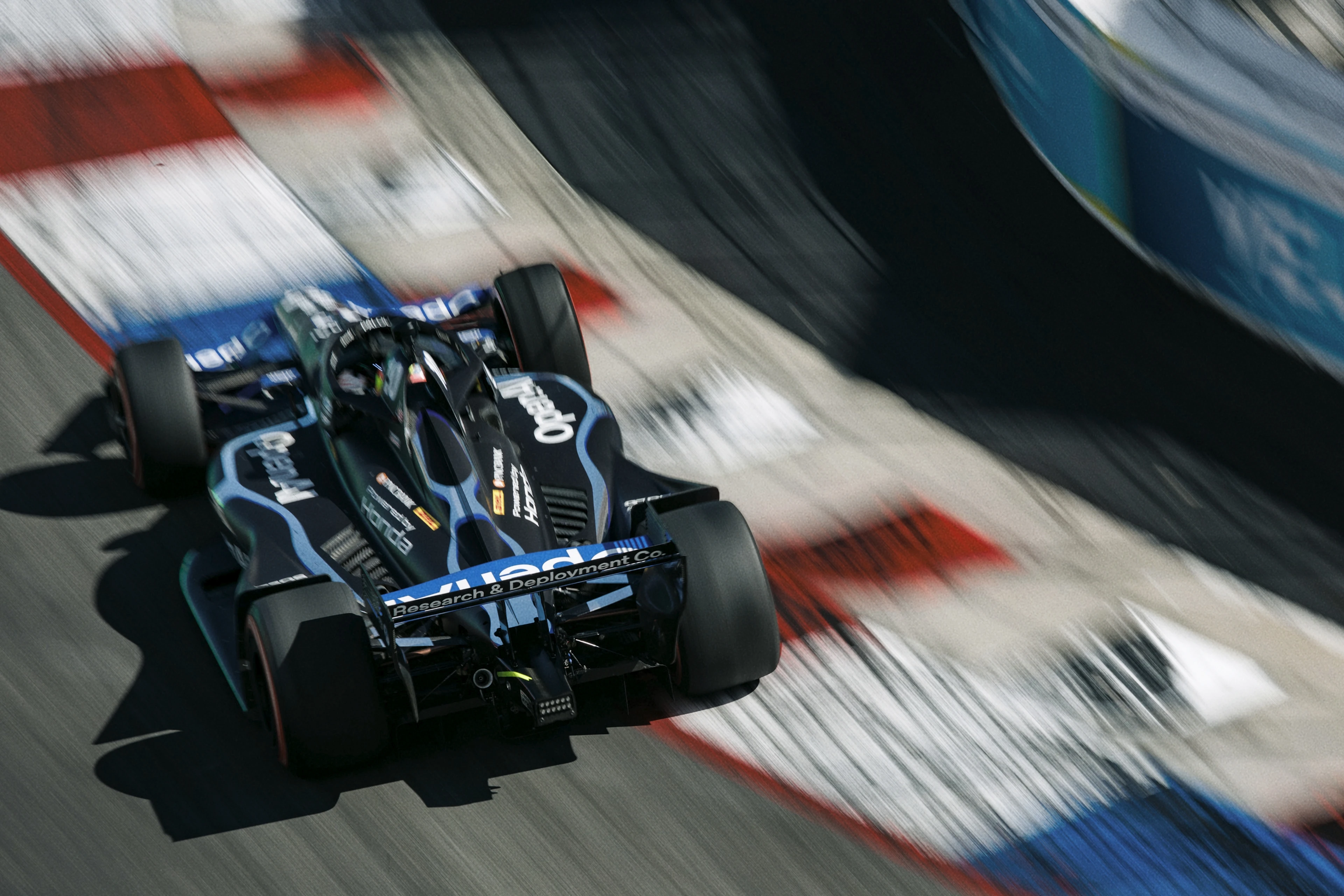A high-speed black and blue Formula 1 race car is captured from behind as it speeds along a racetrack with red, white, and blue painted curbs in bright daylight. The car's rear wing displays the words "Research & Deployment Co." and the image conveys intense motion and blurred surroundings.
