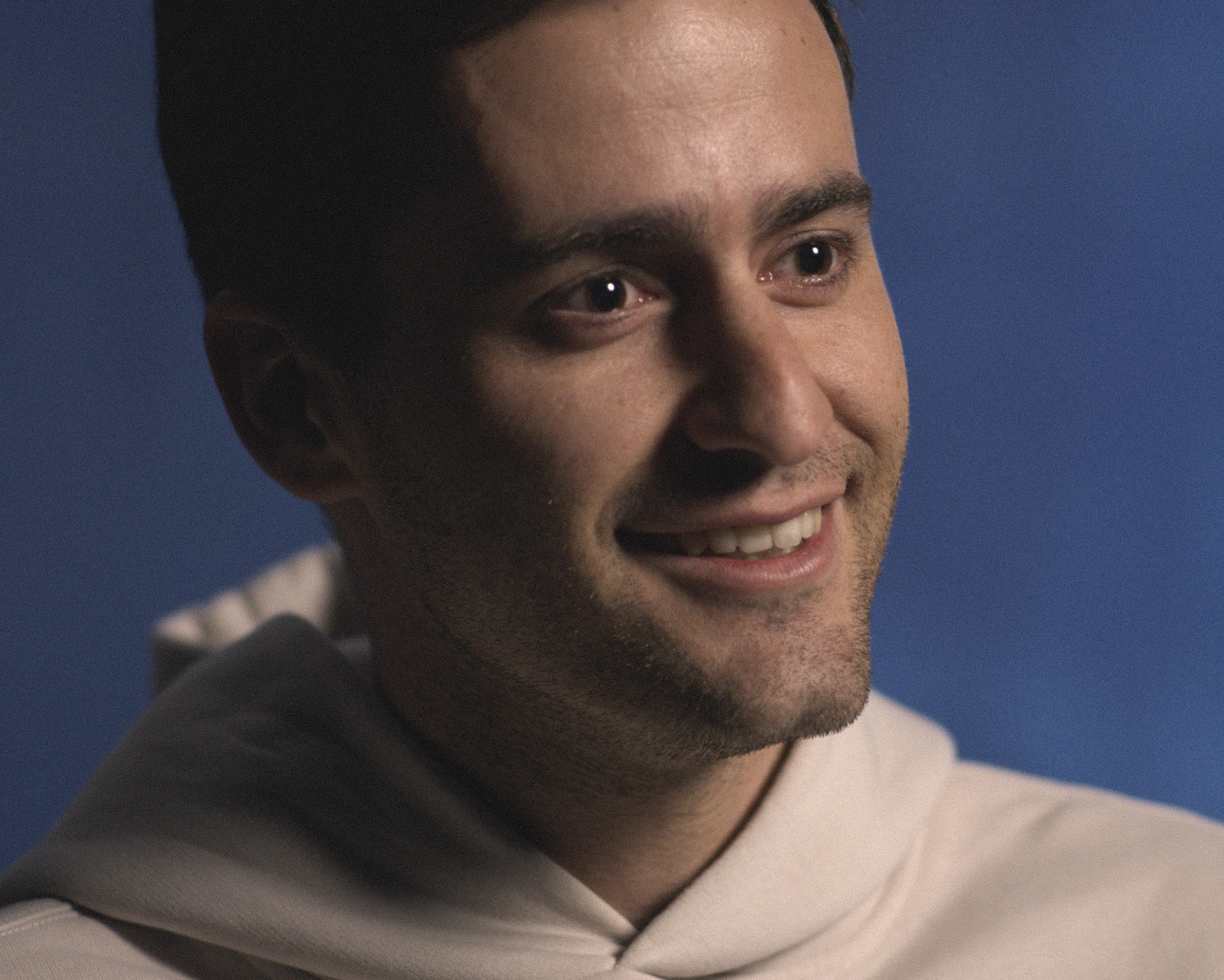 Close-up of a young man with short dark hair wearing a light-colored hoodie, smiling and looking slightly off-camera against a blue background.