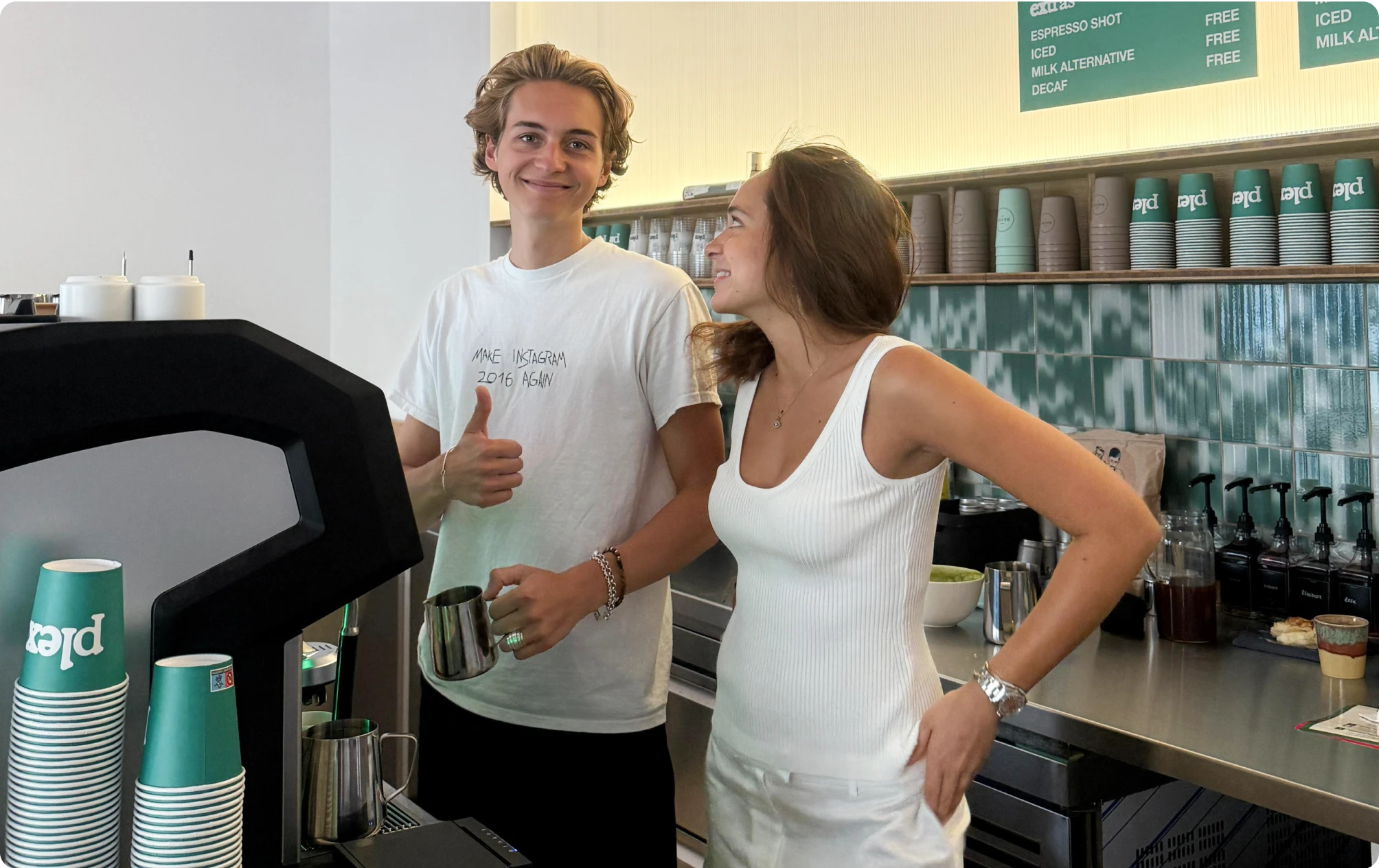 Two people behind a coffee counter smiling. One gives a thumbs up while holding a milk frothing pitcher. Branded “plex” cups line the shelves behind them.