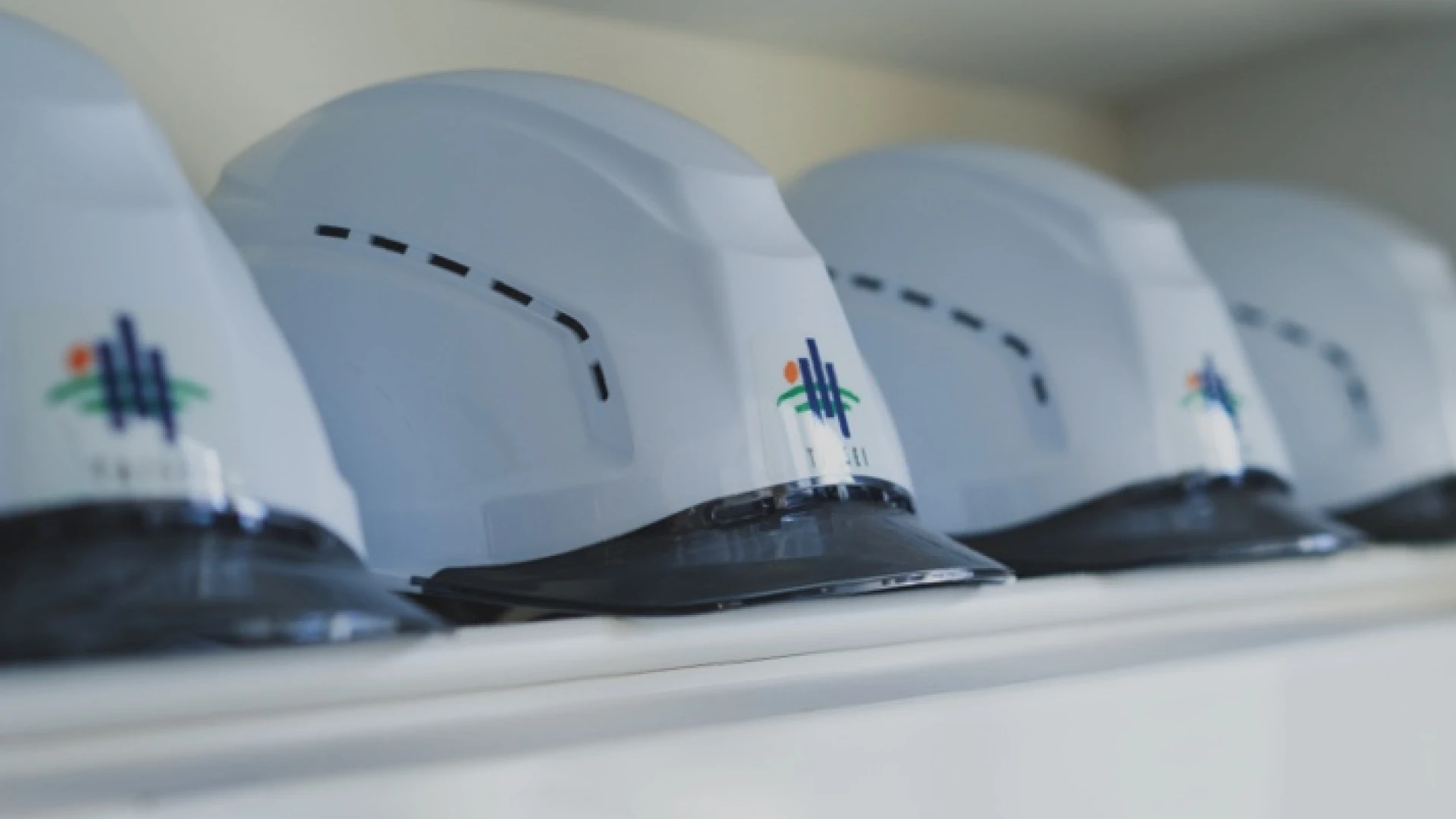 Close-up of several white construction hard hats lined up on a shelf, each with a small multicolored logo, with the nearest helmet in sharp focus and others blurred in the background.