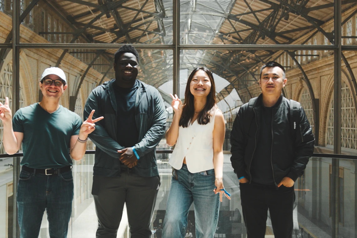 Group photo of four people standing indoors beneath a high arched glass-and-metal ceiling. They are smiling and facing the camera. The person on the far left is wearing glasses, a white cap, and a dark shirt while flashing a peace sign. Next to them, a person in a dark hoodie stands with hands clasped. The third person is wearing a white sleeveless top and jeans, smiling brightly while also flashing a peace sign. The fourth person, on the far right, is wearing a dark jacket with a backpack, standing with hands in pockets.