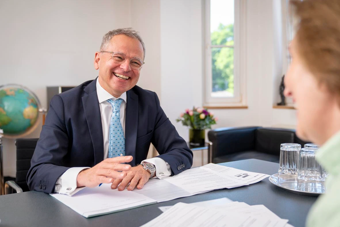 A smiling man in a business suit and light blue tie sits at a desk during a professional consultation. He is holding a pen over a document and appears to be speaking with a client across the table. The setting is a bright, modern office with a globe, flowers, and glasses on a tray in the background.