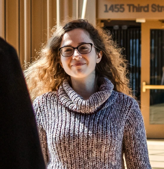 Person wearing glasses and a knit sweater smiling outdoors in front of a building entrance.