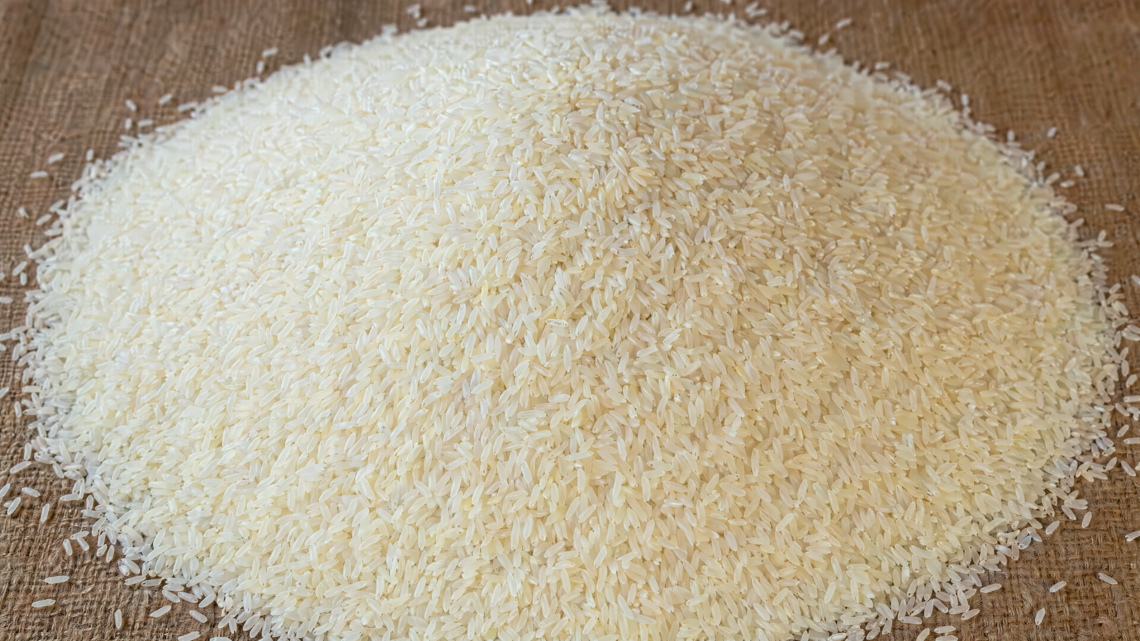 Generated with ChatGPT Images 2.0, this close-up image shows a large mound of uncooked white rice grains piled on a textured burlap surface.