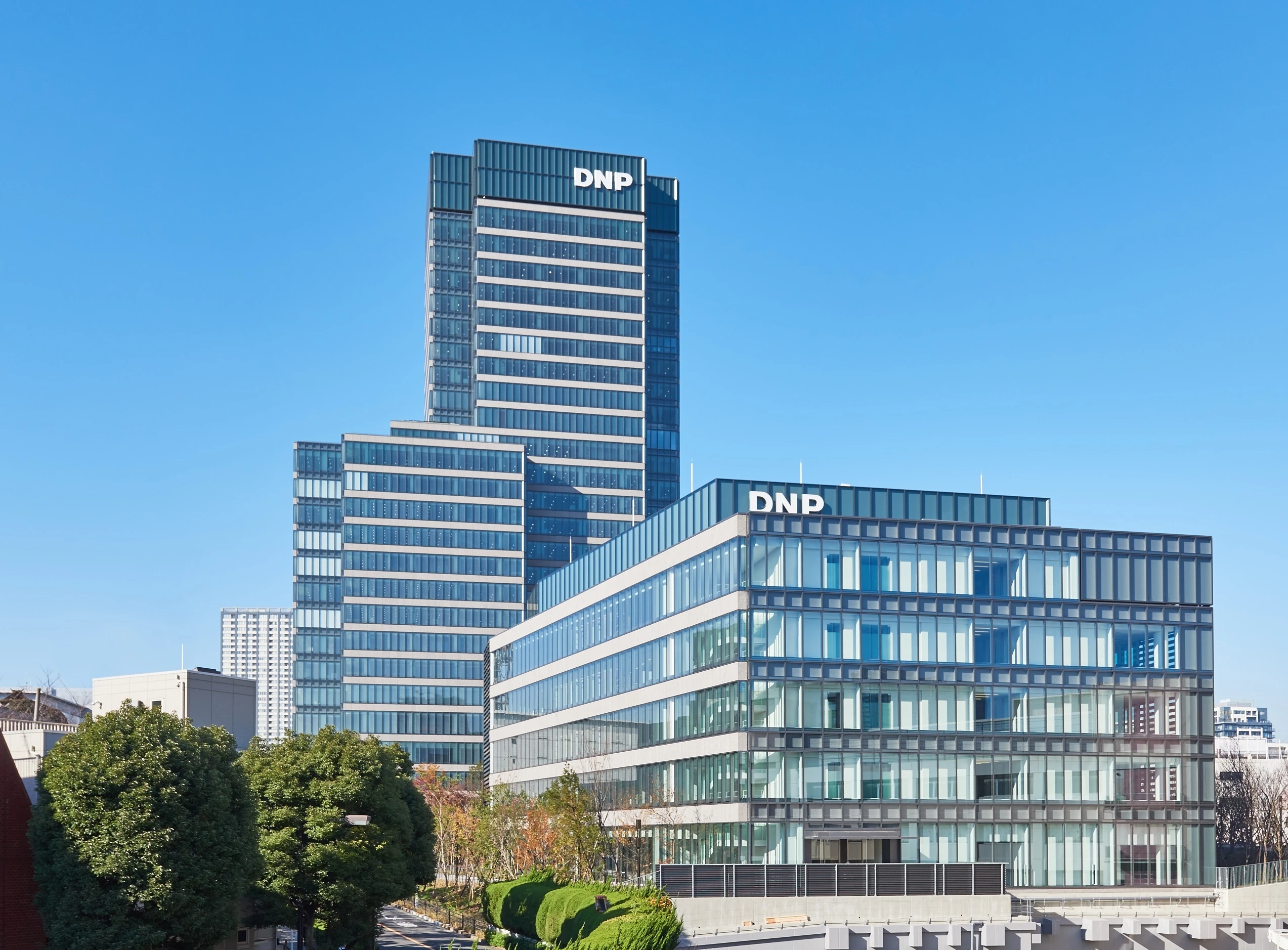 Modern glass office buildings with “DNP” logos under a clear blue sky, surrounded by trees and city structures.
