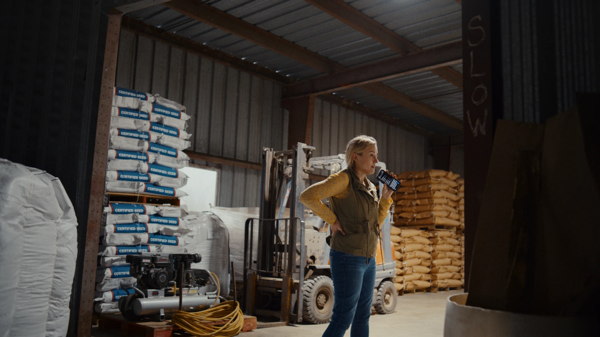 A woman stands inside a farm storage building, speaking into a handheld radio beside stacked bags of certified seed and a forklift.