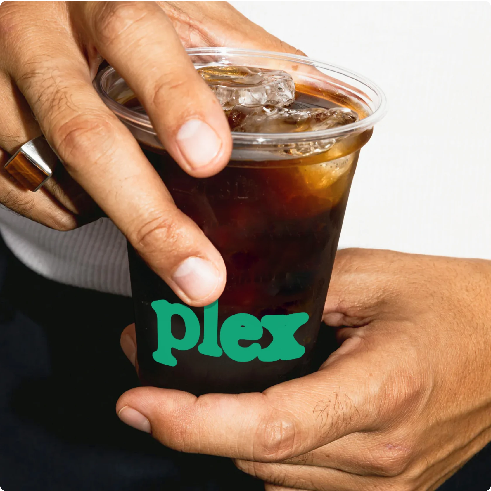 Close-up of a person holding a plastic cup of iced coffee with the word “plex” in green lettering on the cup. The person is wearing a ring and gripping the lid.