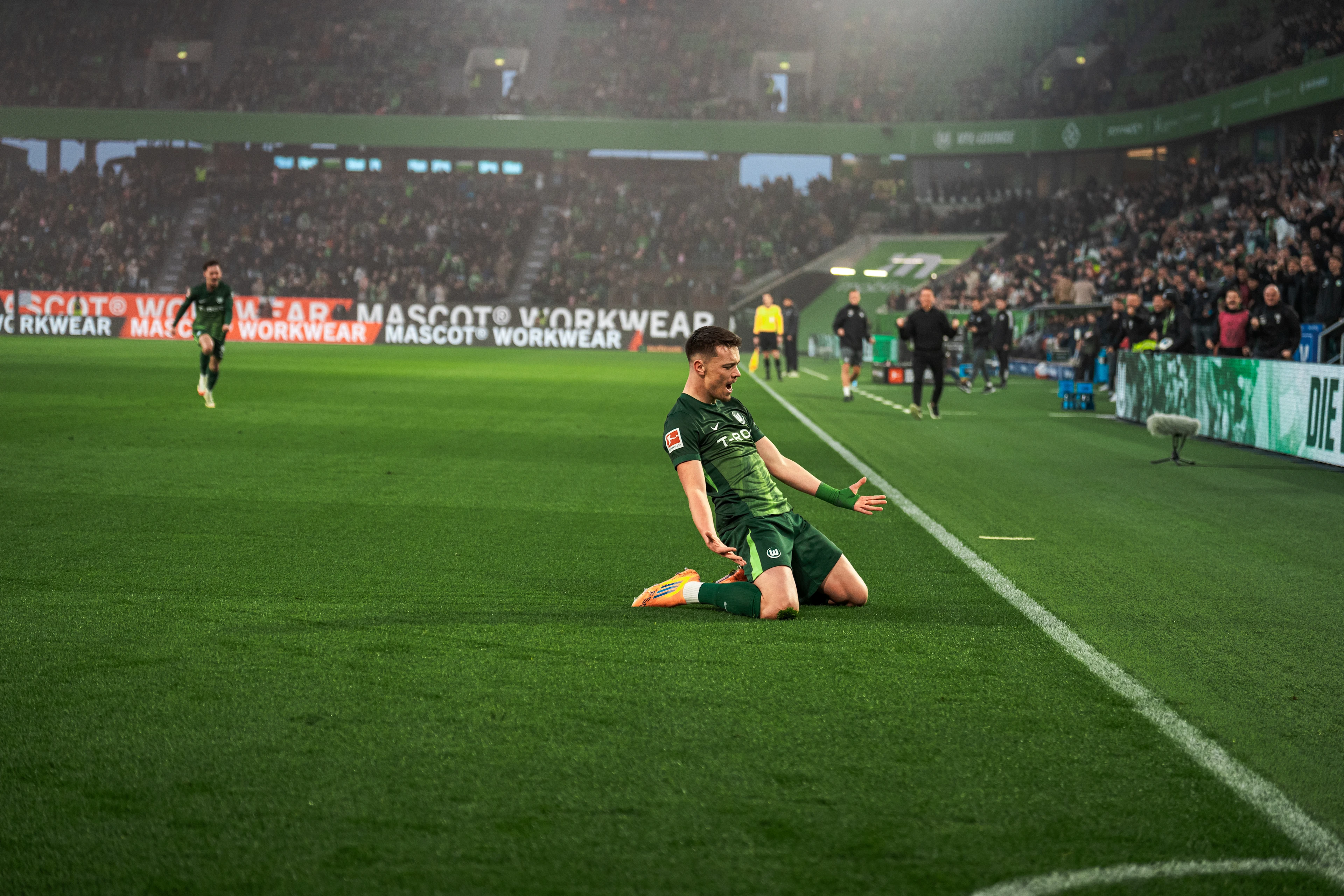 VfL Wolfsburg player slides on his knees in celebration after scoring, with teammates and fans in the background.