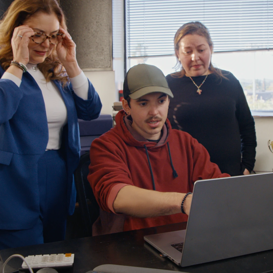A young man works on a laptop while two women stand beside him, watching the screen and discussing the work in an office setting.