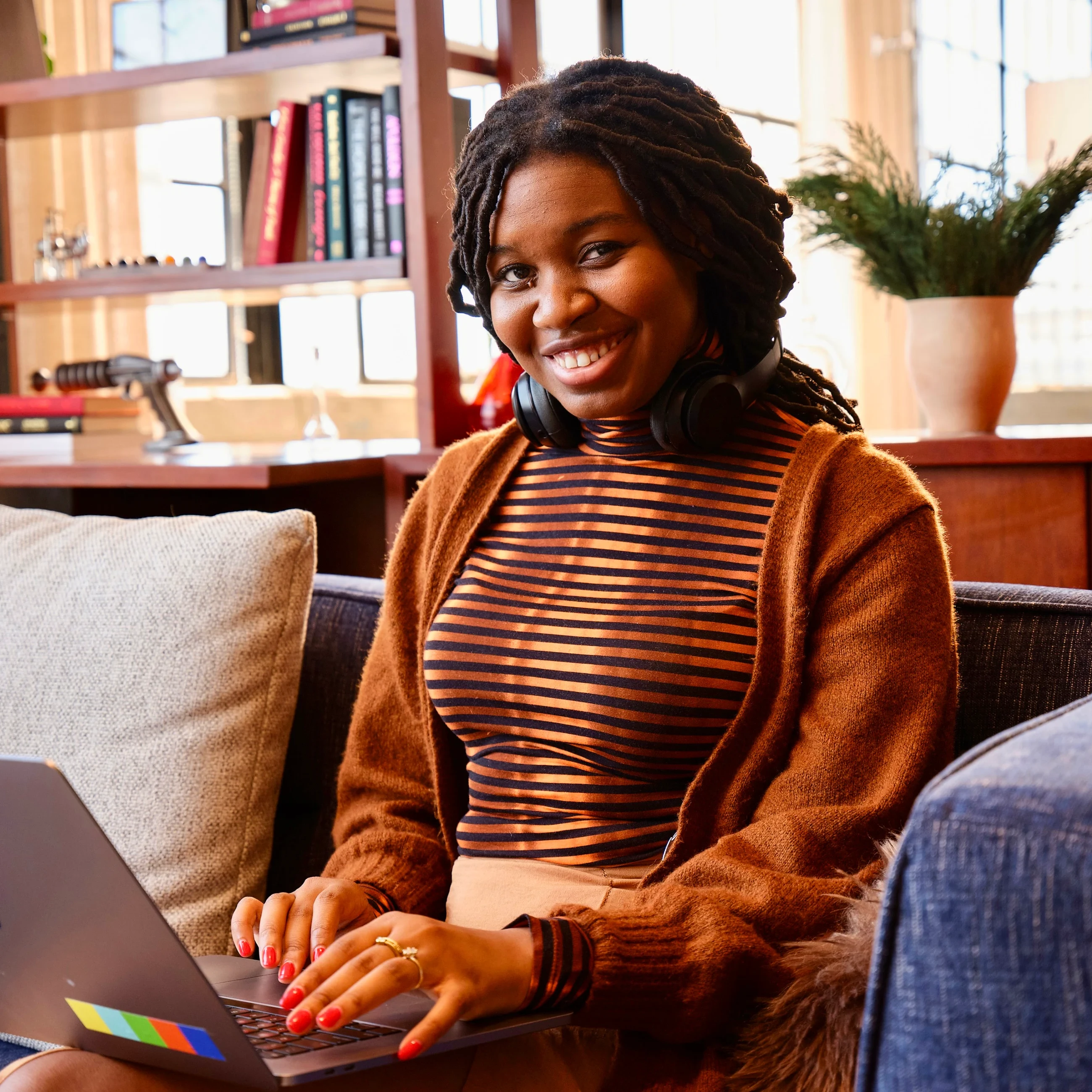 A smiling woman with headphones around her neck, working on a laptop in a cozy room with a bookshelf and a plant in the background. She's wearing a striped top and a brown cardigan.