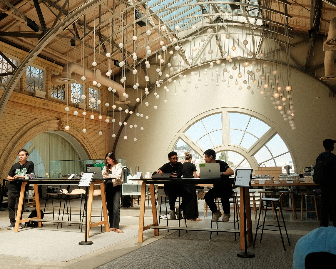 Bright, airy indoor space with high arched ceilings, hanging lights, and a large circular window letting in natural light. Several people are seated or standing at tall tables labeled “Build Bar,” working on laptops or writing. The atmosphere resembles a collaborative workspace or event setup, with modern furniture and an open, inviting design.