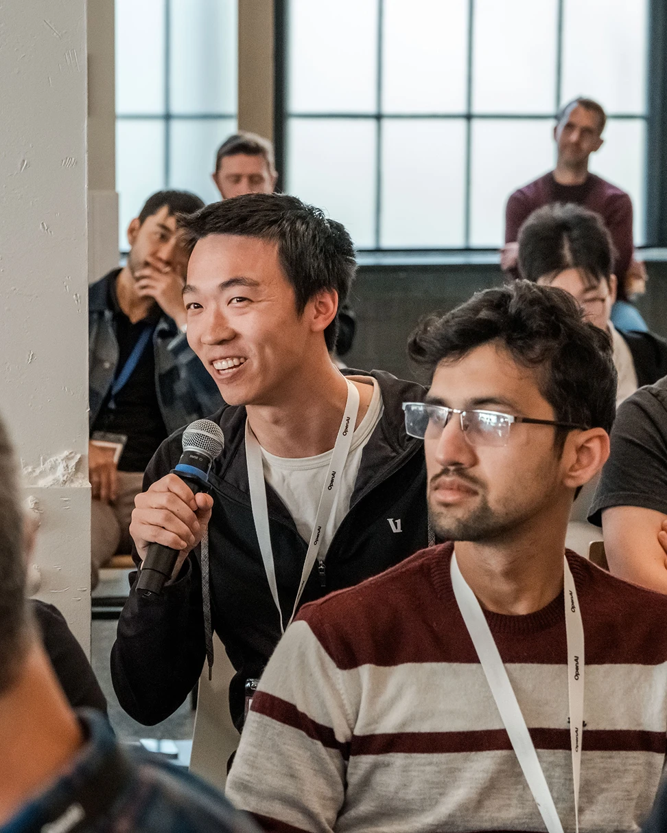 A person is smiling while holding a microphone and speaking during an event. They are wearing a black jacket and a white lanyard with “OpenAI” text on it. In the foreground, another attendee in glasses and a striped sweater is listening attentively. Several more people are seated in the background, with large windows letting in natural light.