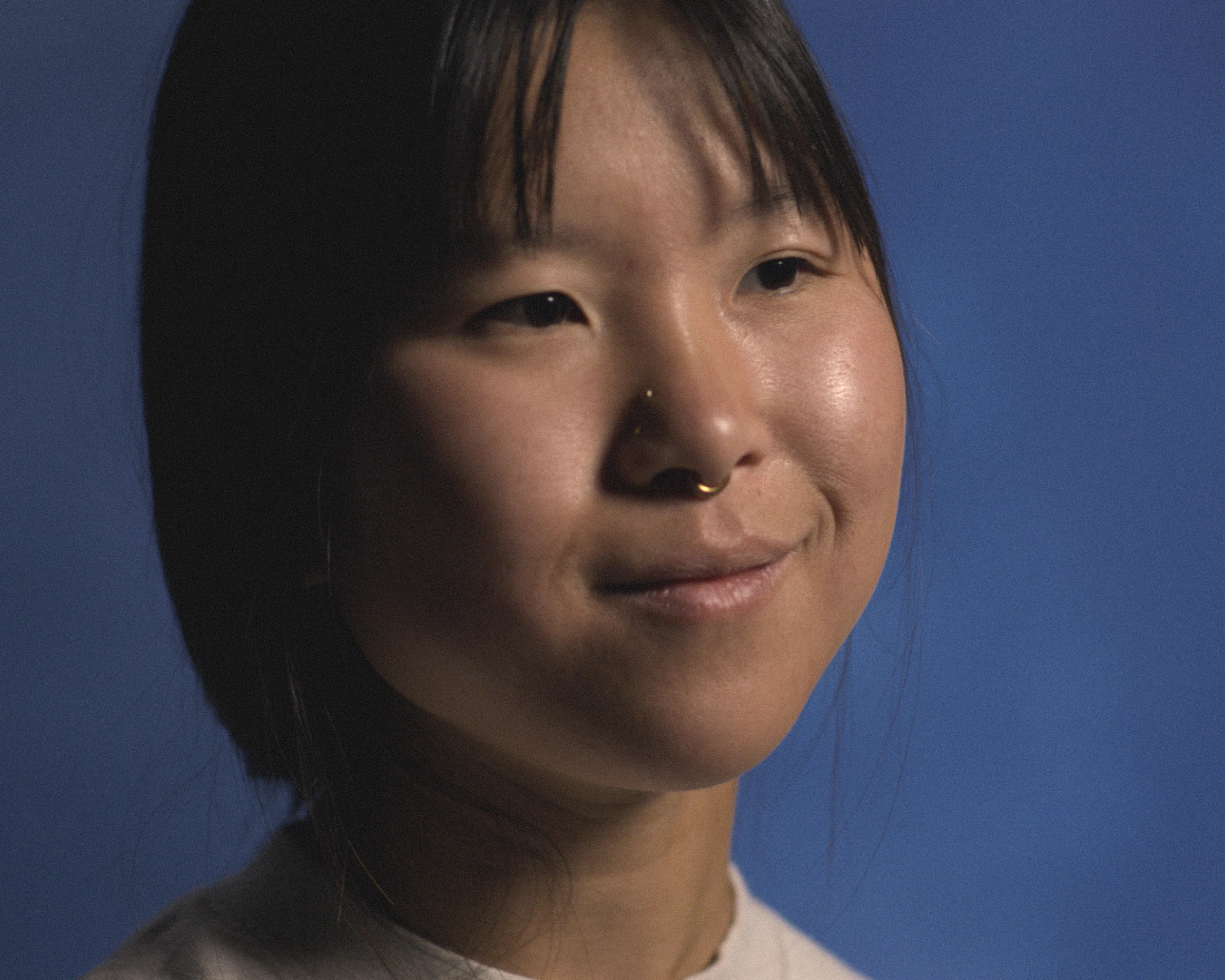 Close-up portrait of a person with medium-length dark hair, a nose ring, and a slight smile, against a plain blue background.