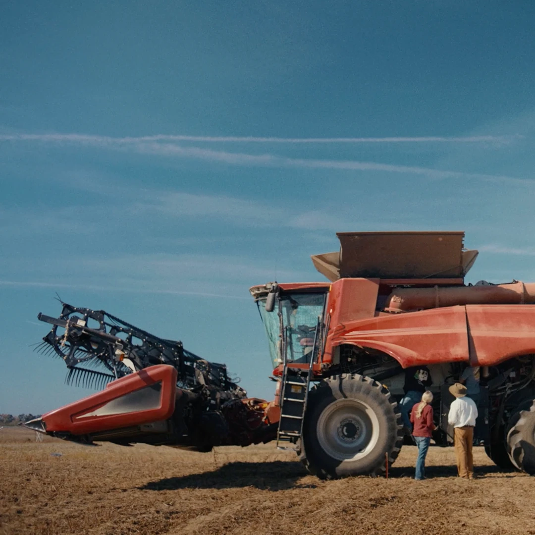 Two people stand beside a large red combine harvester in an open, harvested field under a wide blue sky.