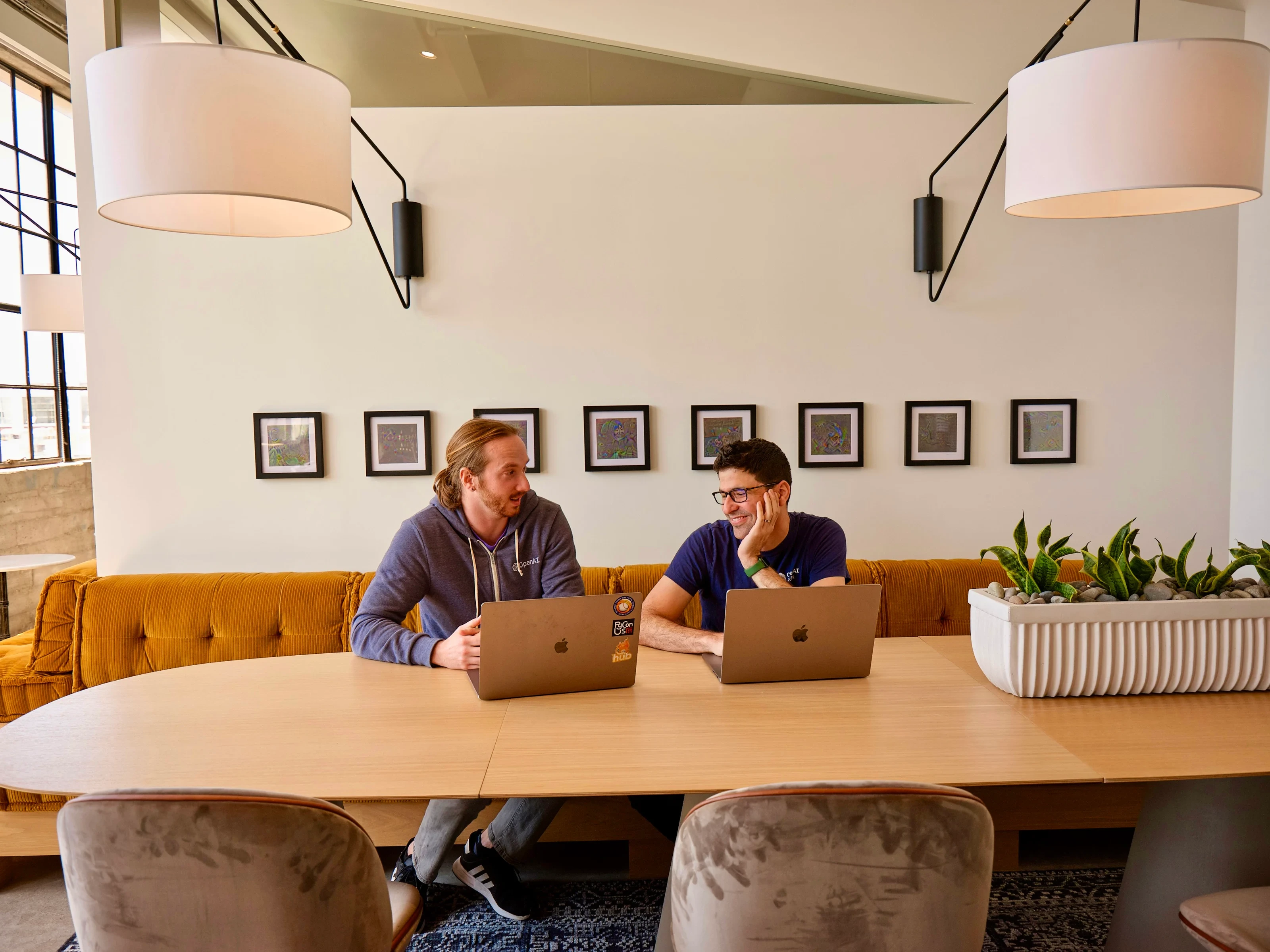  Two men smiling at each other, seated at a table with laptops in a well-lit, modern room with a mustard sofa, framed pictures on the wall, indoor plants, and pendant lights.