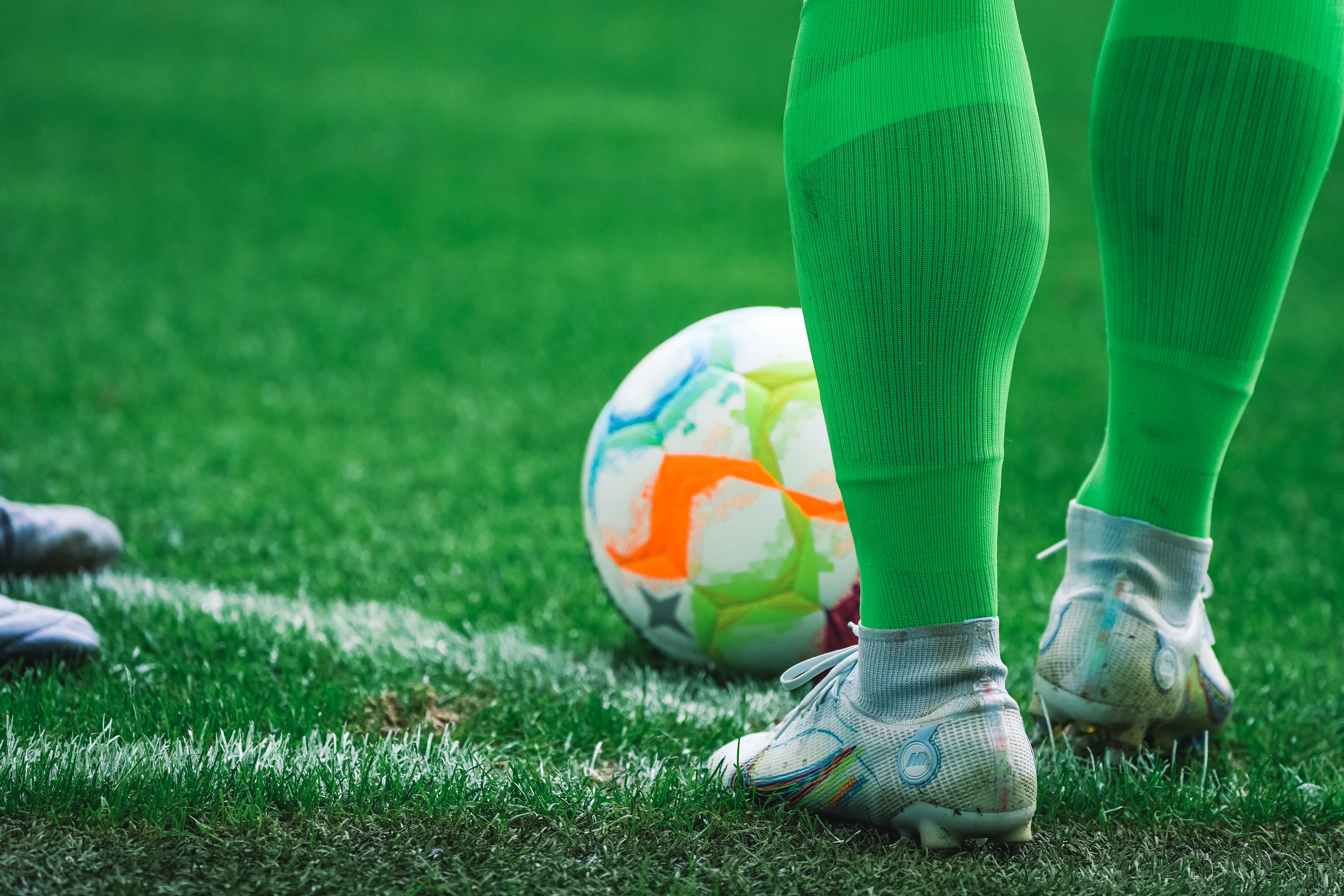 Close-up of a soccer ball on the grass near a player’s green socks and white cleats, poised for a kickoff or free kick.