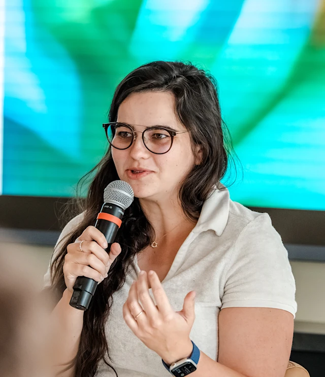 A person with long dark hair and glasses speaks into a handheld microphone while gesturing with their other hand. They are wearing a light-colored collared shirt and a smartwatch. The background is blurred with bright colors, including green and blue tones.