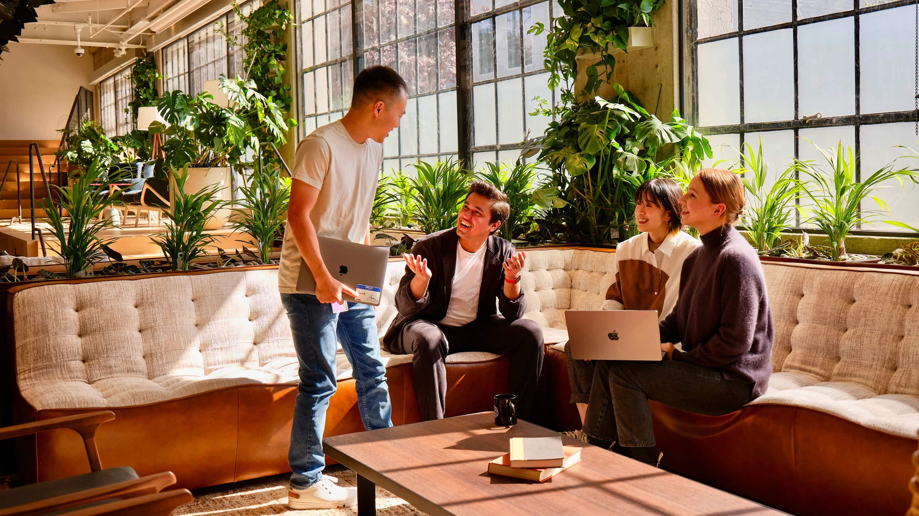 明亮的、摆满植物的办公室，有大窗户，四个人正在讨论；其中三个人坐着，手里拿着笔记本电脑，一个人站着，手里拿着笔记本电脑。