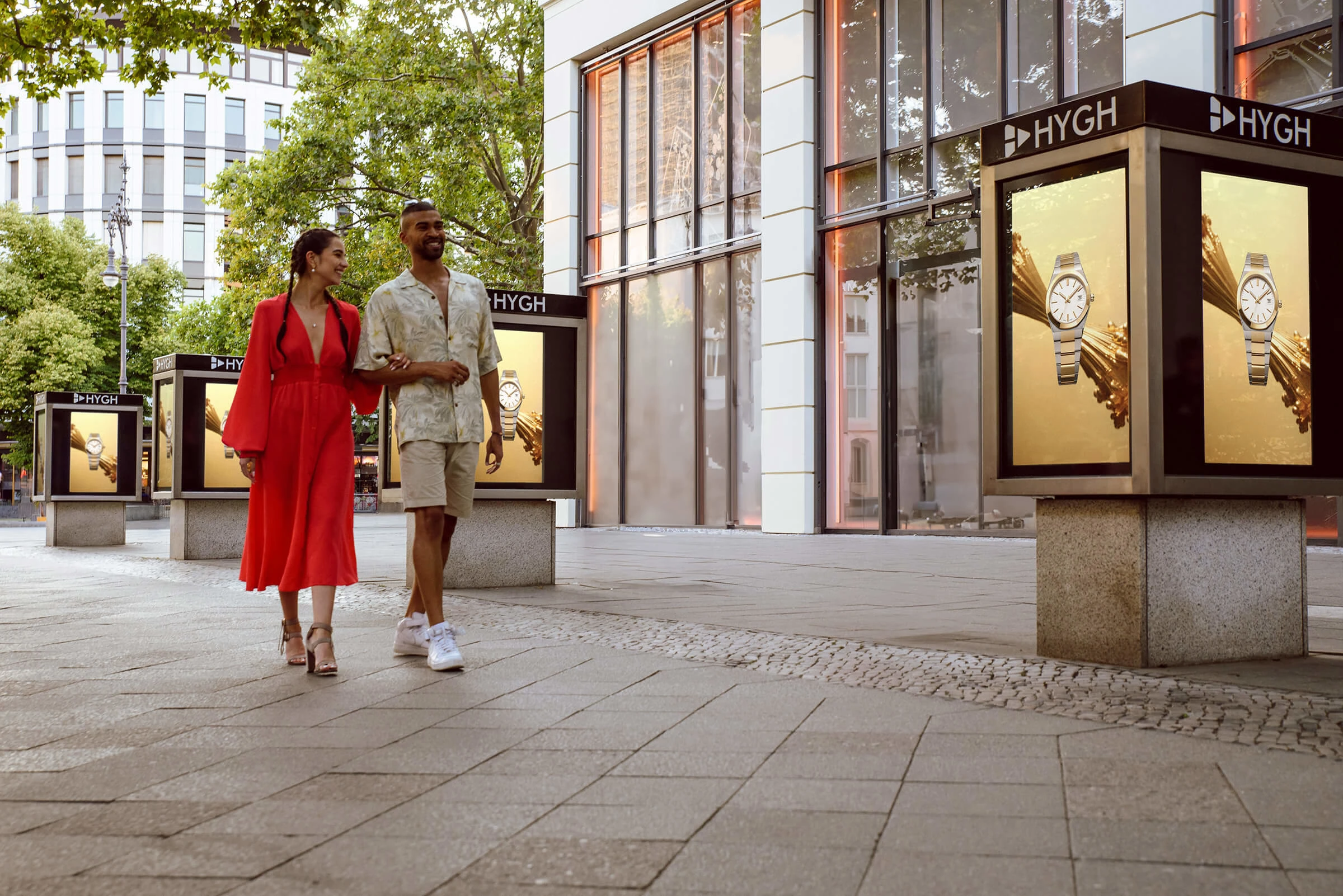 Two people walk arm-in-arm past a series of HYGH-branded digital displays on a sidewalk. The screens show luxury watch ads. The setting is a modern urban shopping district.