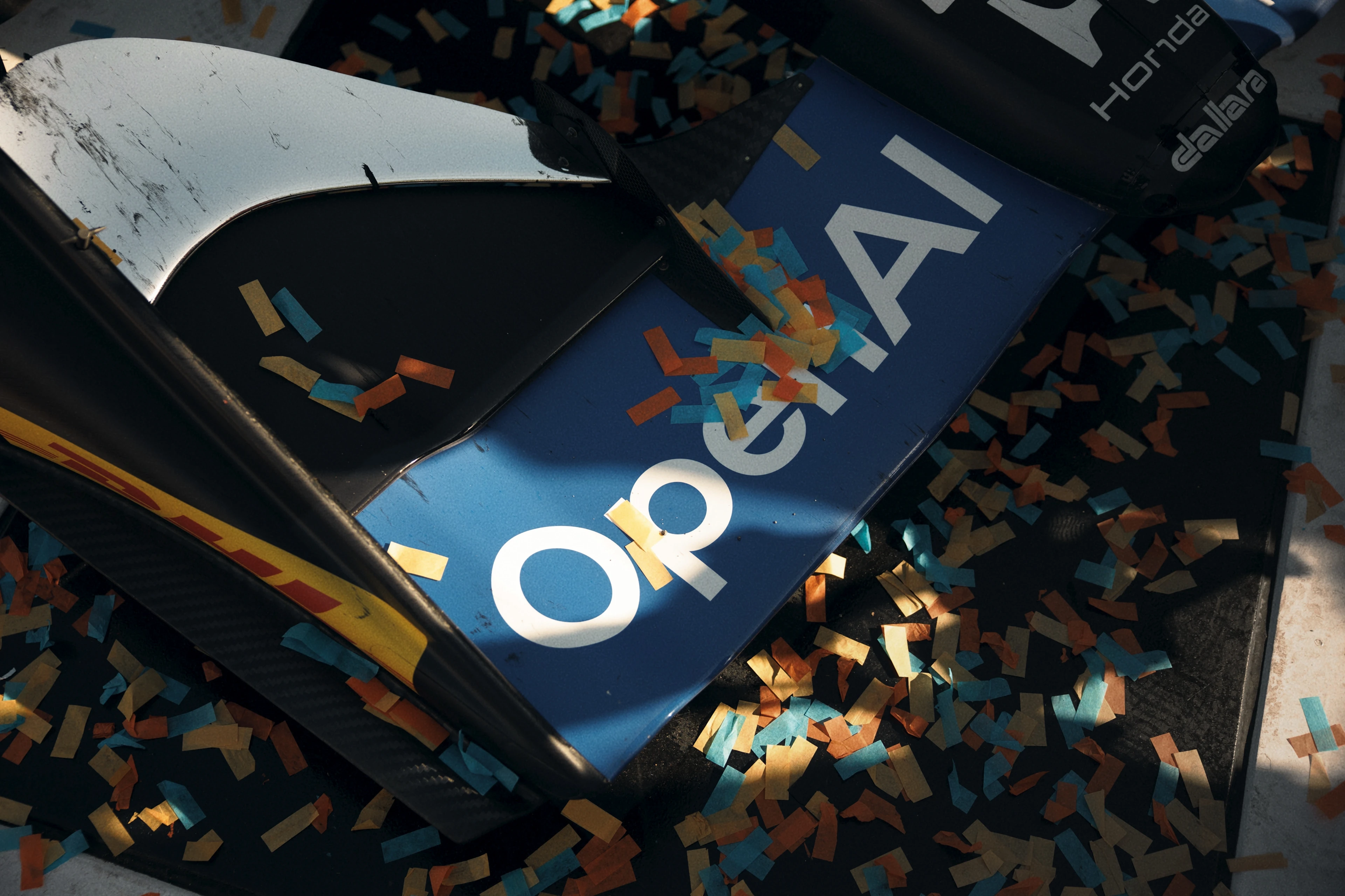 Close-up of a blue and black racing car wing with the word "OPENAI" printed on it, scattered with multicolored confetti in yellow, blue, and orange.