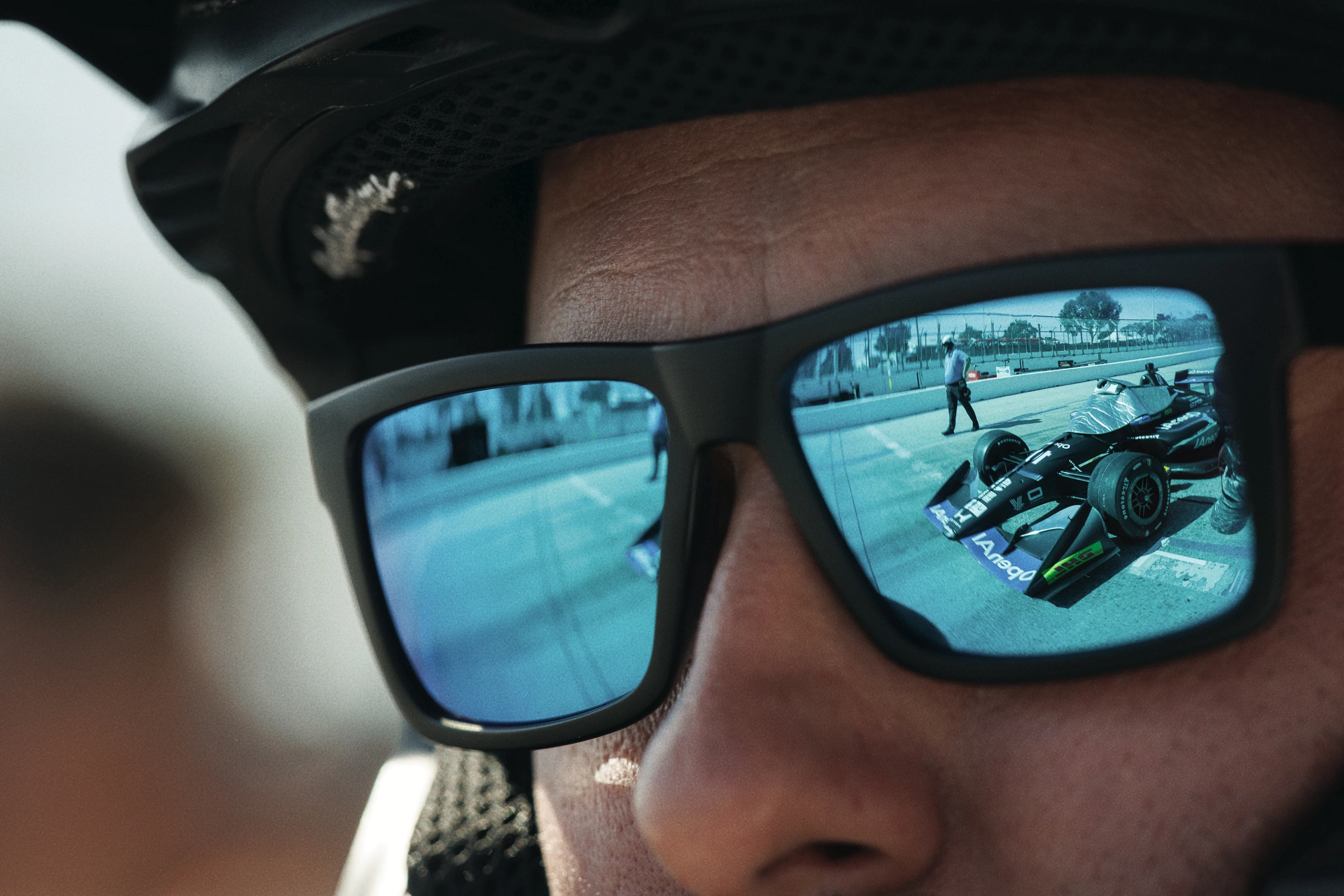 Close-up of a person wearing sunglasses and a helmet, with a reflection of a black race car and a person in a white shirt on a track visible in the lenses.