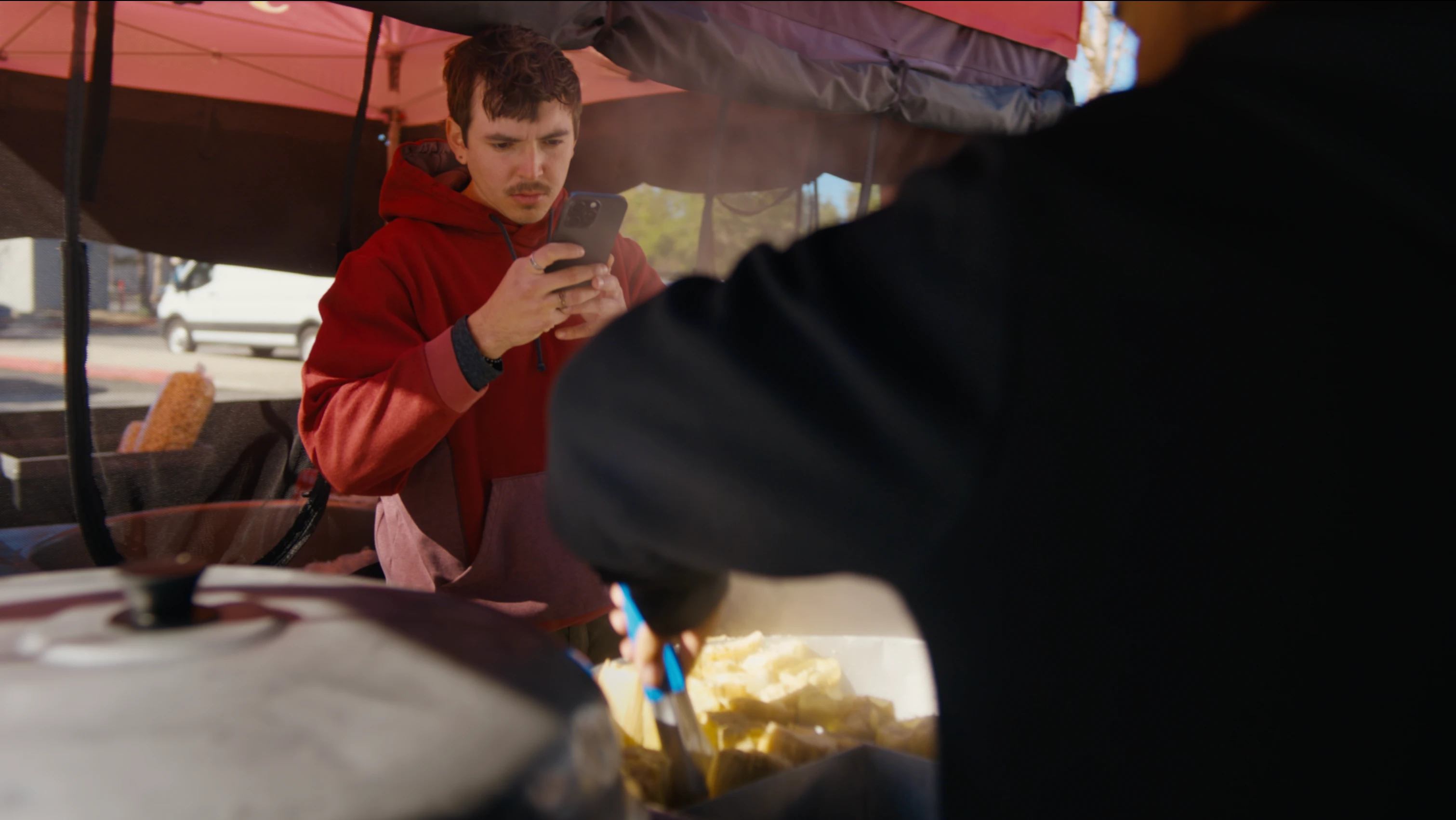A young man in a red hoodie films food with his smartphone at a street food stand while another person prepares food in the foreground.