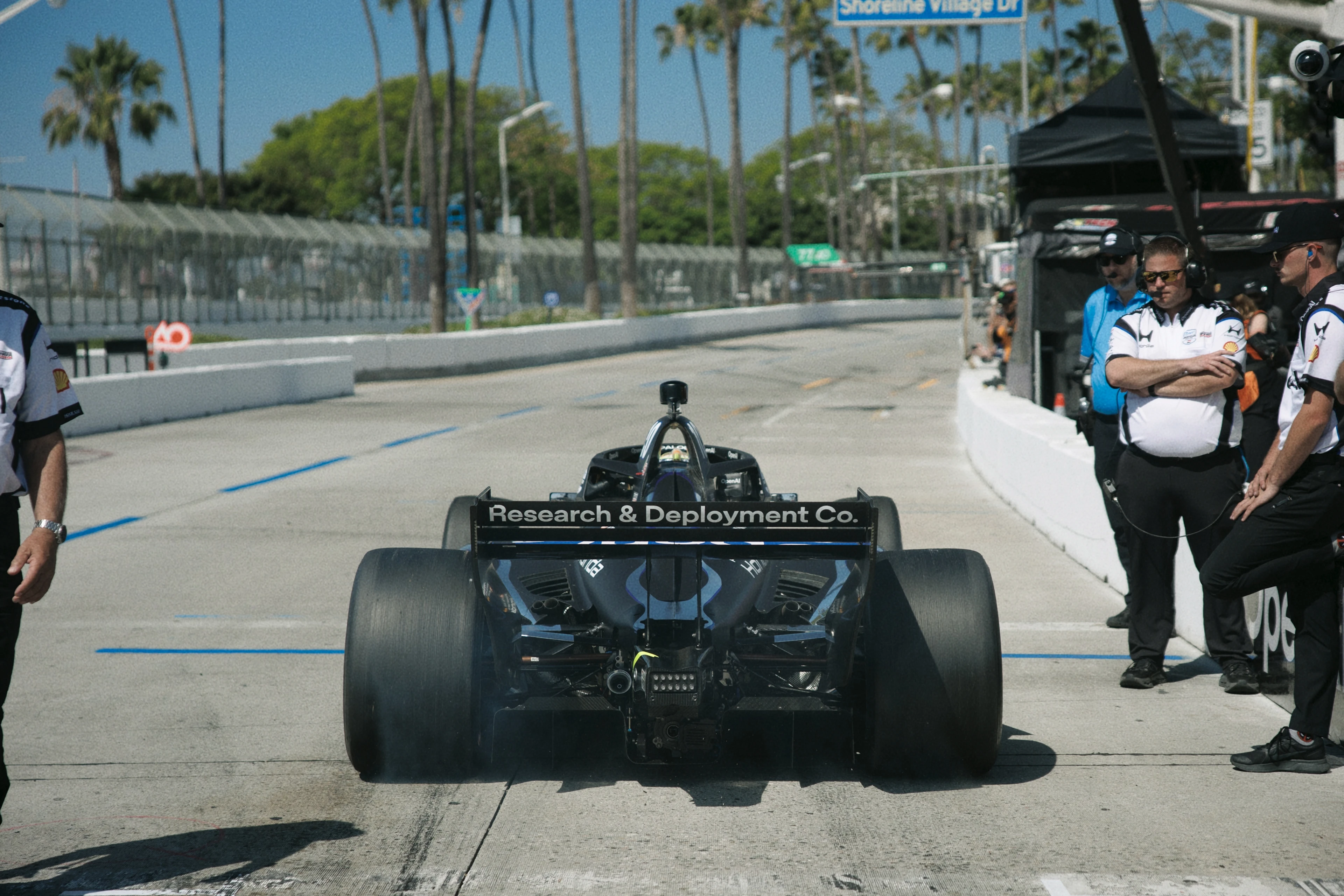 Rear view of a black race car in a pit lane with "Research & Deployment Co." on its rear wing, surrounded by crew members in white and black uniforms.