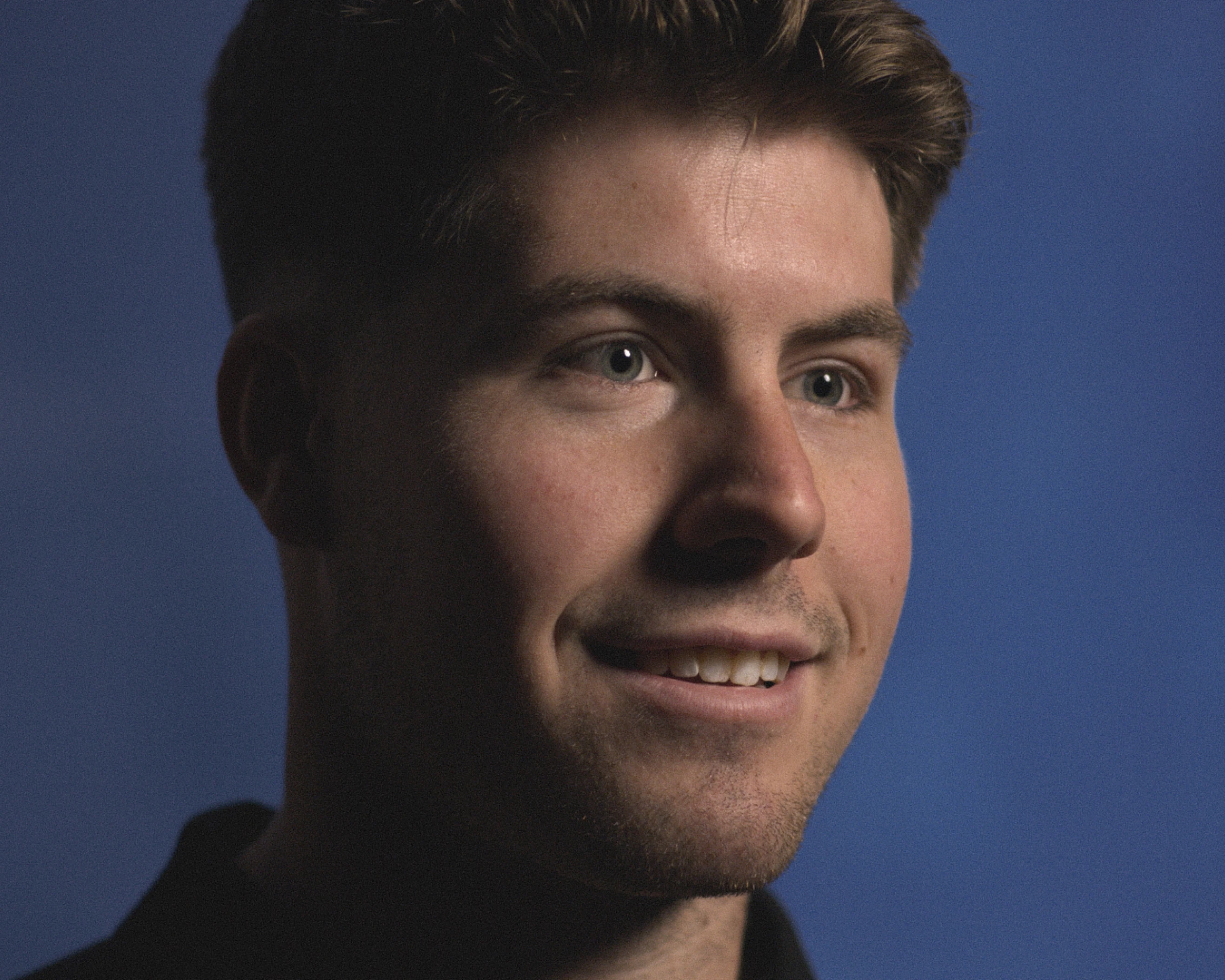 Close-up portrait of a young man with short brown hair, light stubble, and green eyes, smiling slightly against a plain blue background.