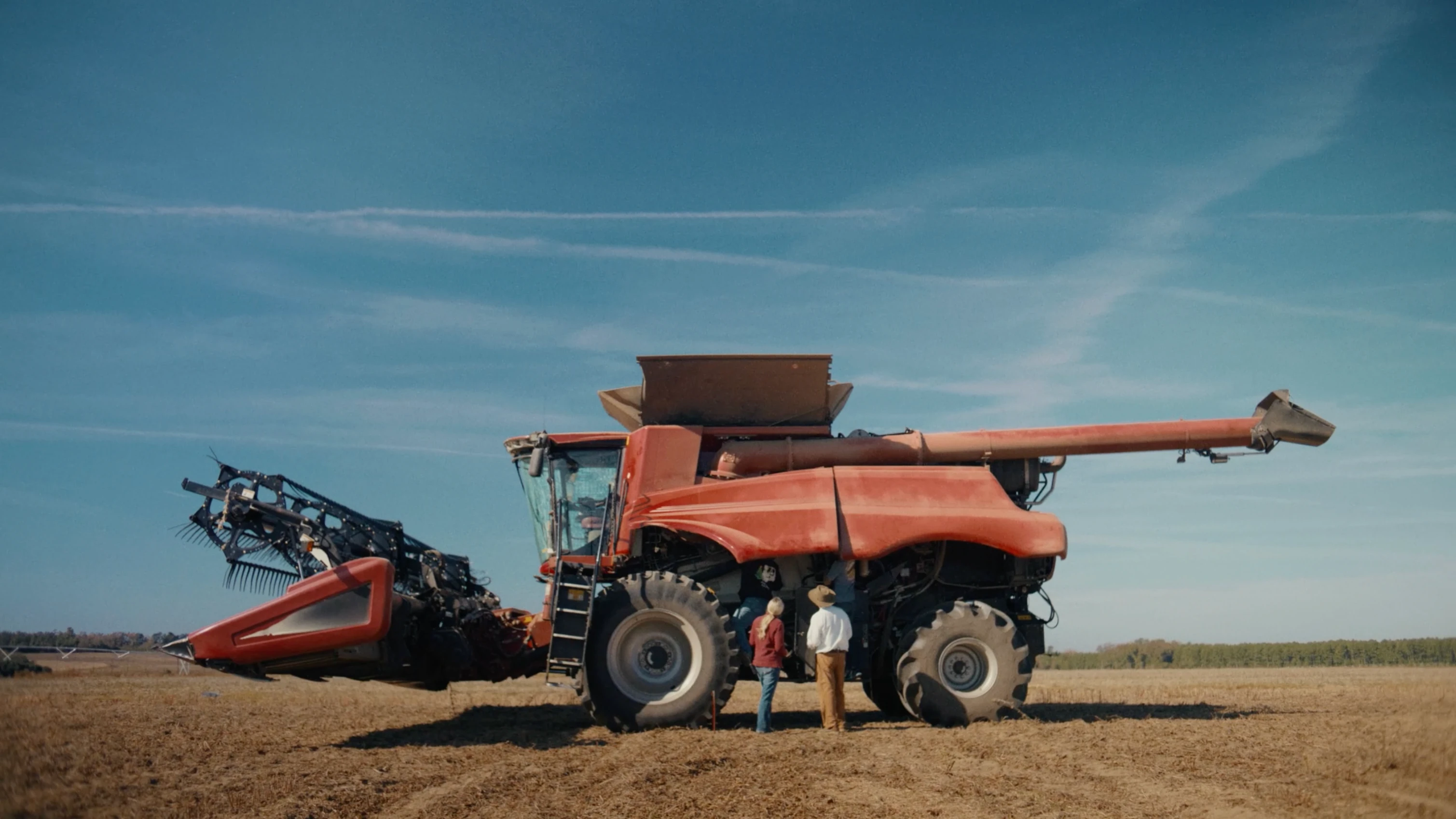 Two people stand beside a large red combine harvester in an open, harvested field under a wide blue sky.