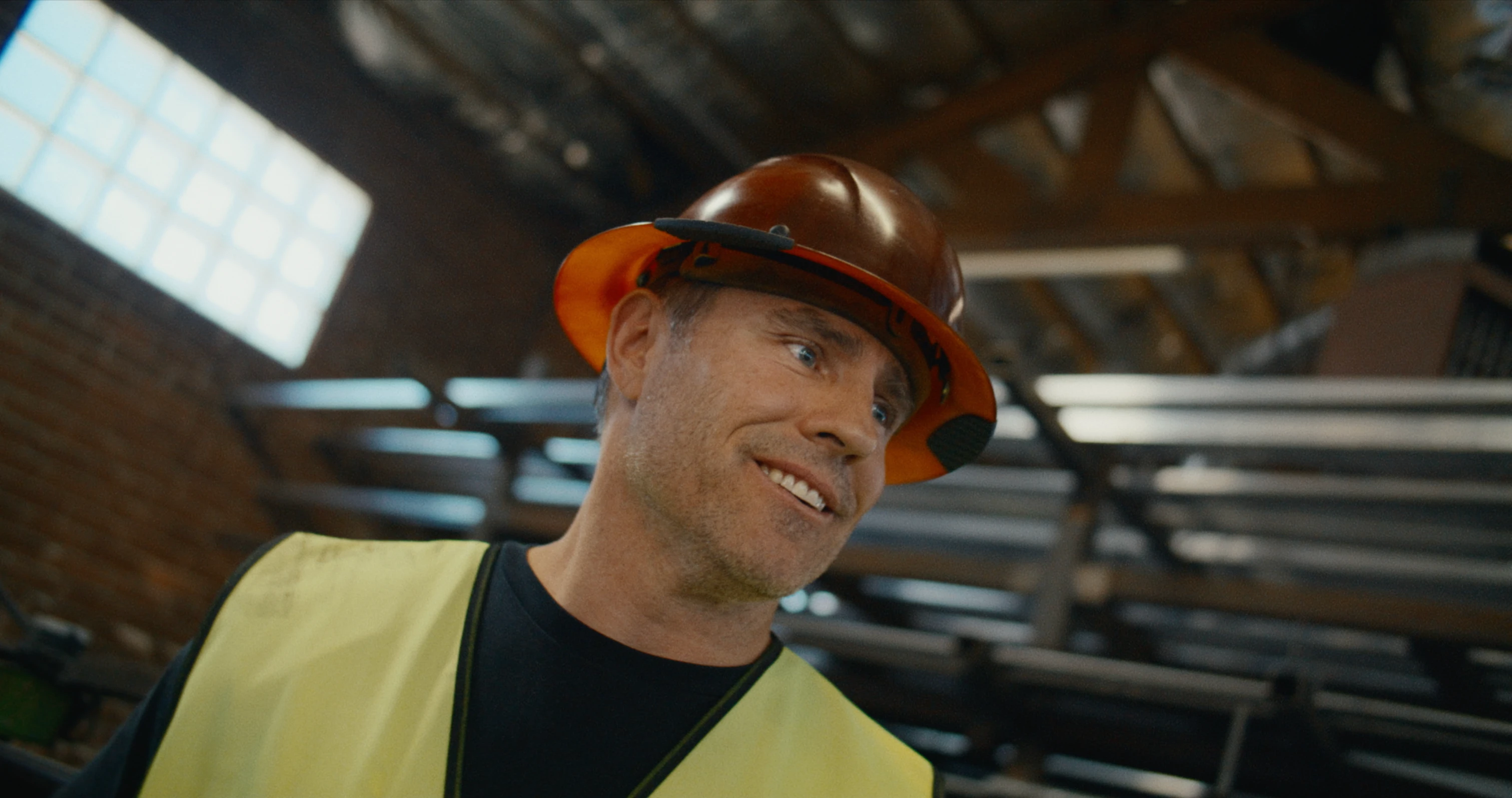 A smiling worker wearing a hard hat and yellow safety vest stands inside an industrial workshop with metal beams in the background.