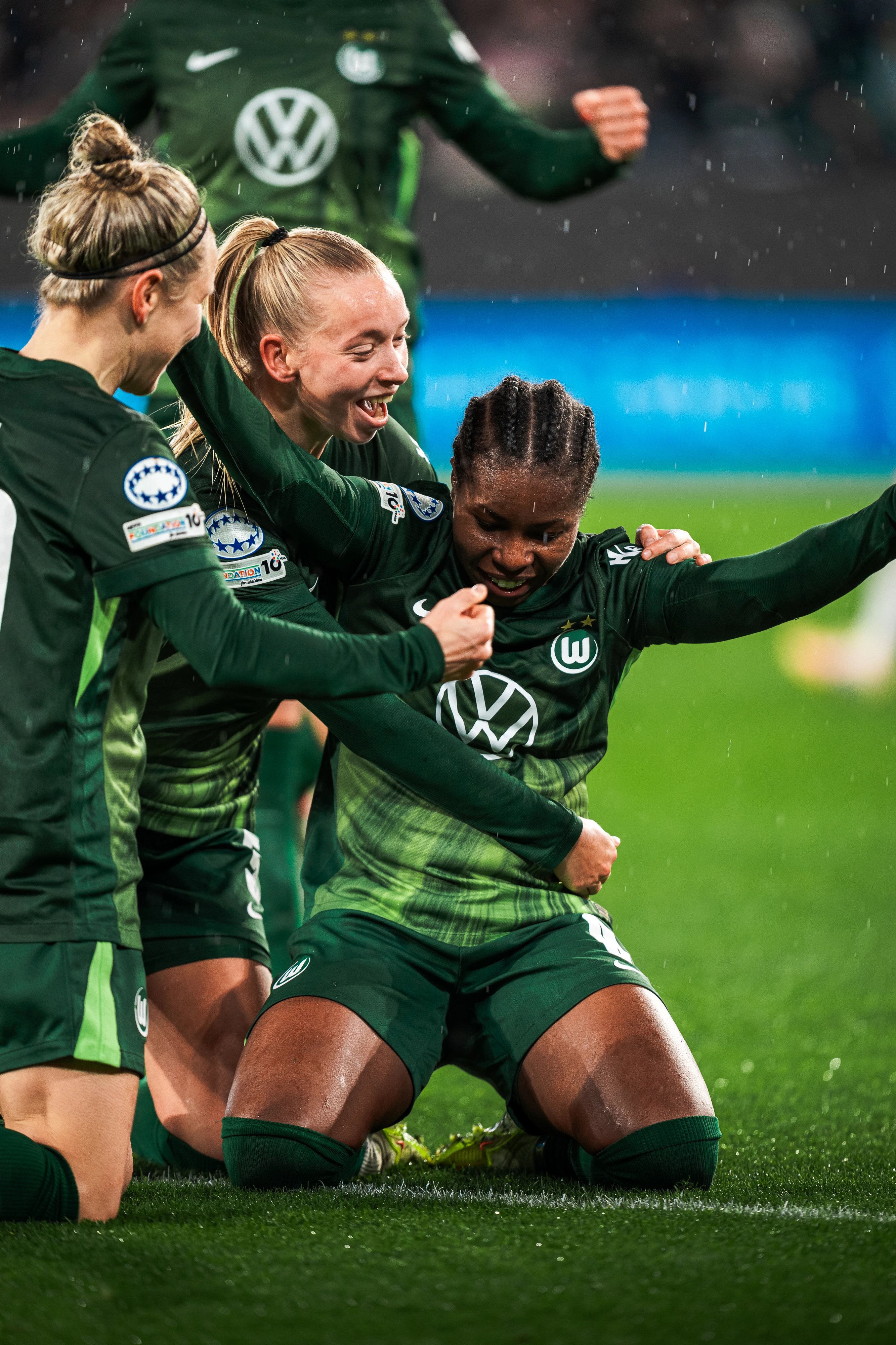 VfL Wolfsburg women’s players celebrate a goal together on the pitch in the rain, wearing green kits.
