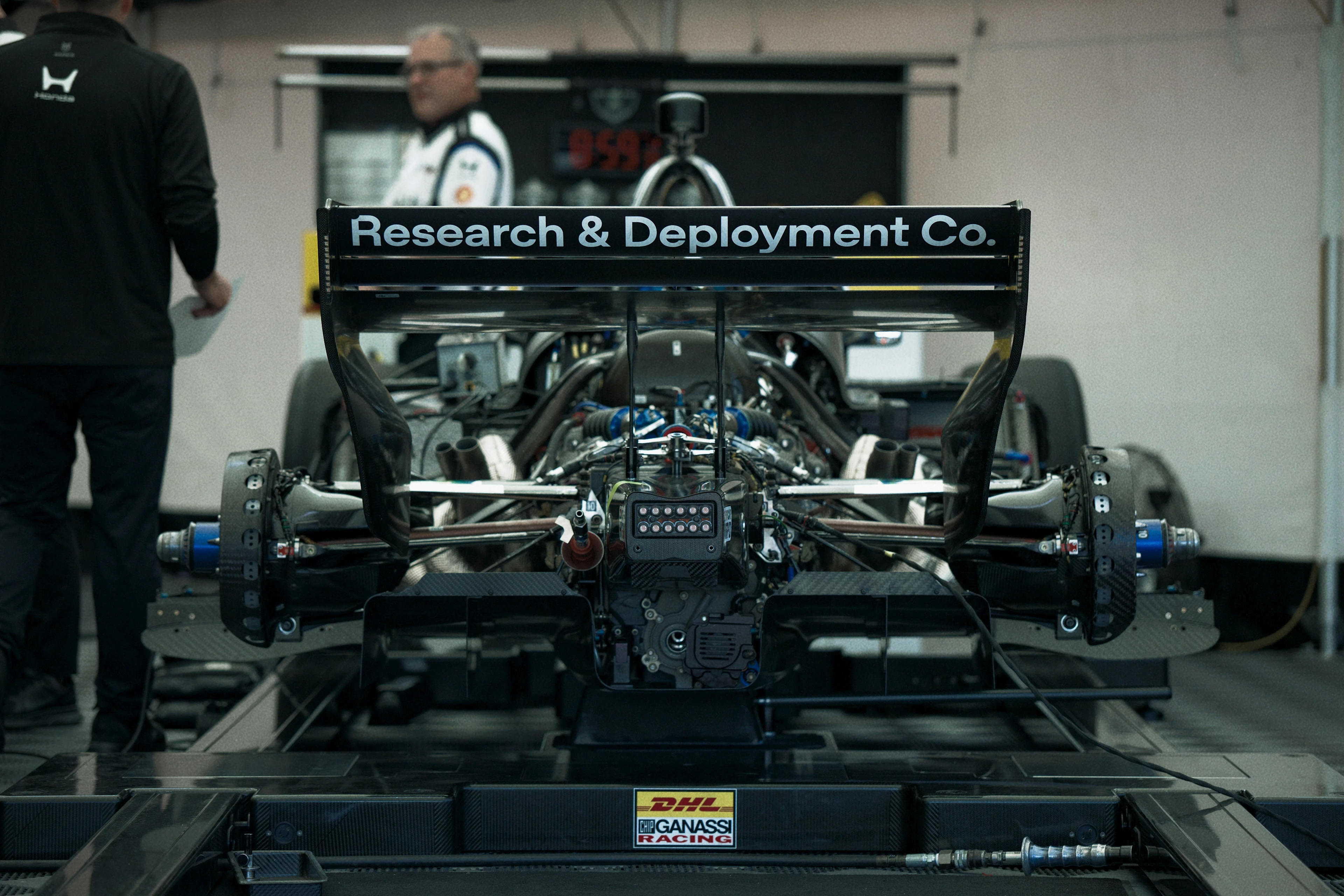 Rear view of a race car in a garage with its rear wing displaying "Research & Deployment Co." Two people stand nearby, one in a black jacket and the other in a white racing uniform. The car has visible mechanical components and a DHL Chip Ganassi Racing logo on its rear diffuser.