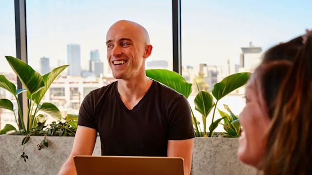 A man sitting at a table with a laptop, smiling during a conversation in a modern office with large windows. Plants and city buildings are visible in the background, and another person is partially visible in the foreground.