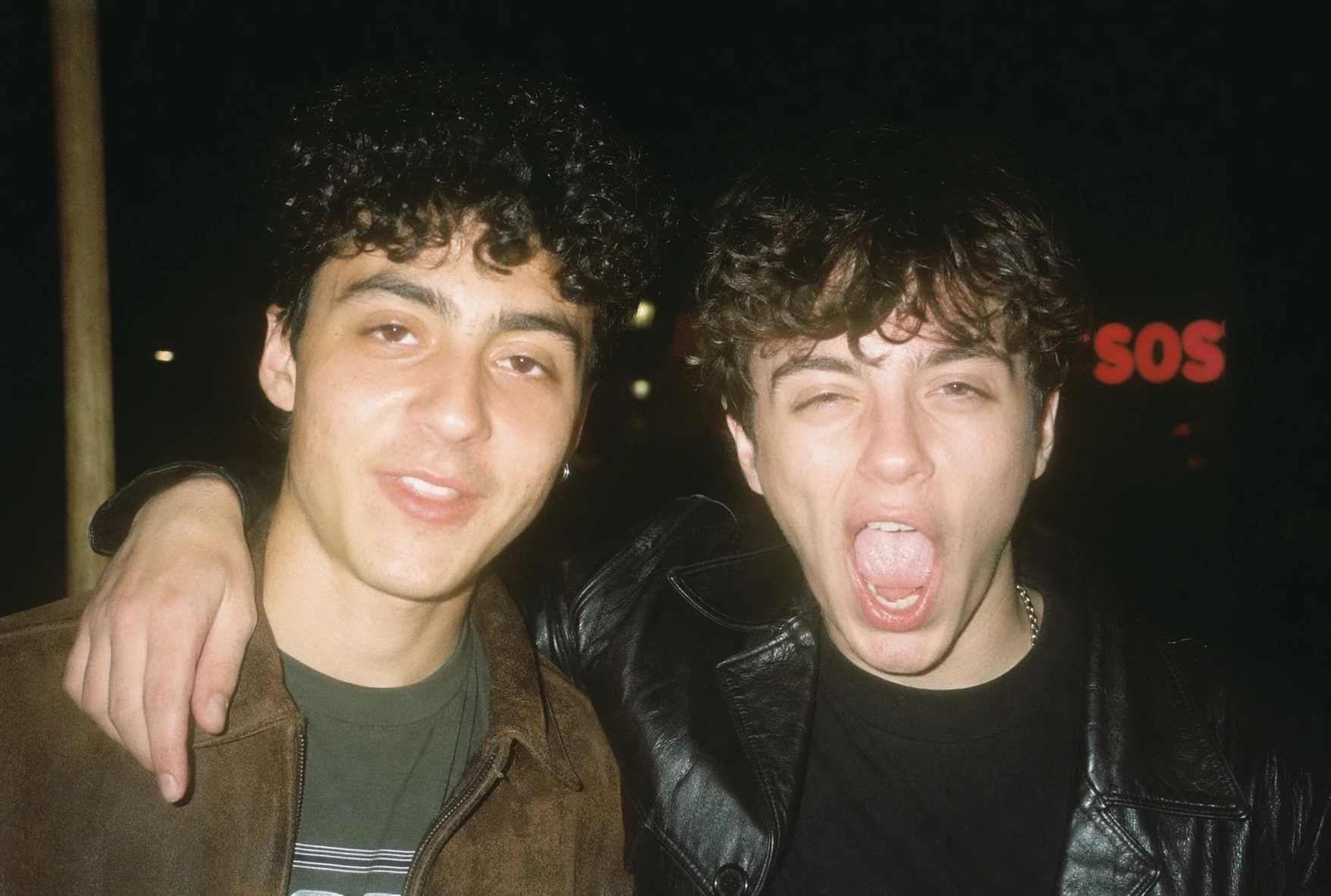 Generated with ChatGPT Images 2.0, this candid nighttime flash photo shows two friends posing closely together on a city street, one smiling at the camera while the other shouts playfully, creating a spontaneous film-camera party snapshot.