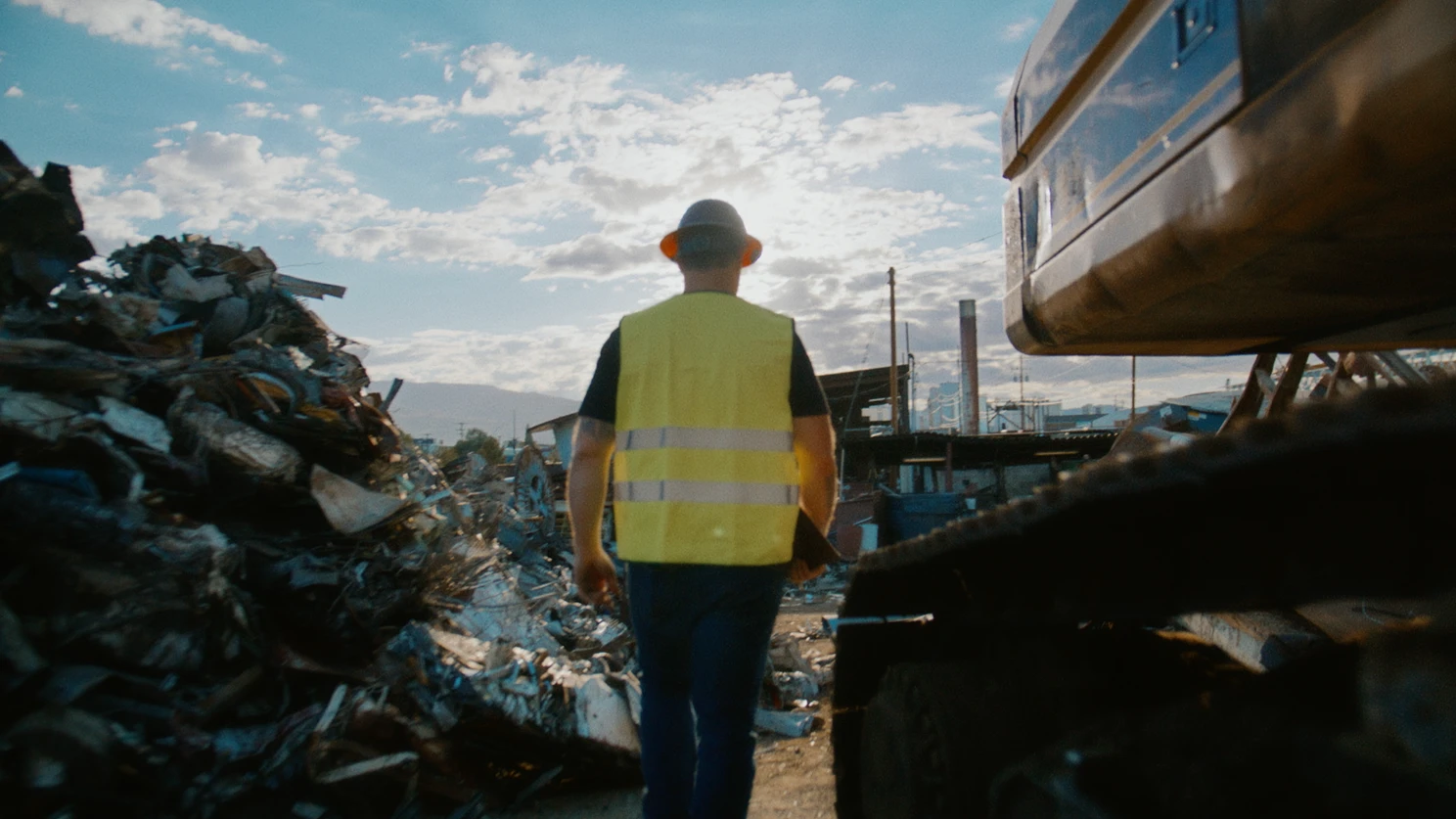 A worker wearing a yellow high-visibility vest and hard hat walks through a scrapyard between piles of metal debris and heavy machinery under a partly cloudy sky.