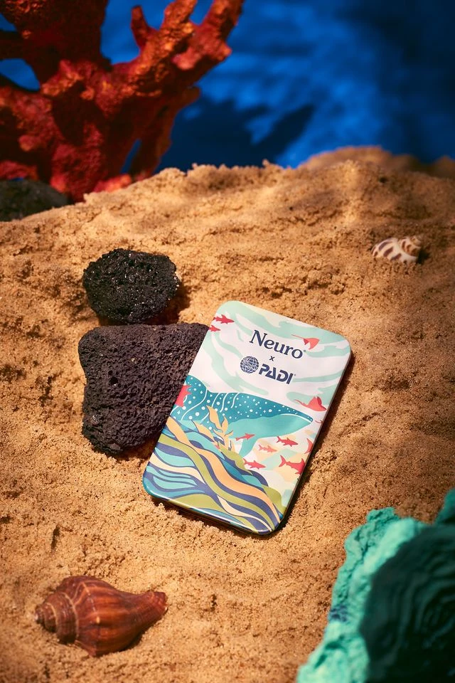 Tin of Neuro x PADI mints placed on sand surrounded by coral, rocks, and seashells, designed with an ocean-inspired illustration featuring fish and waves.