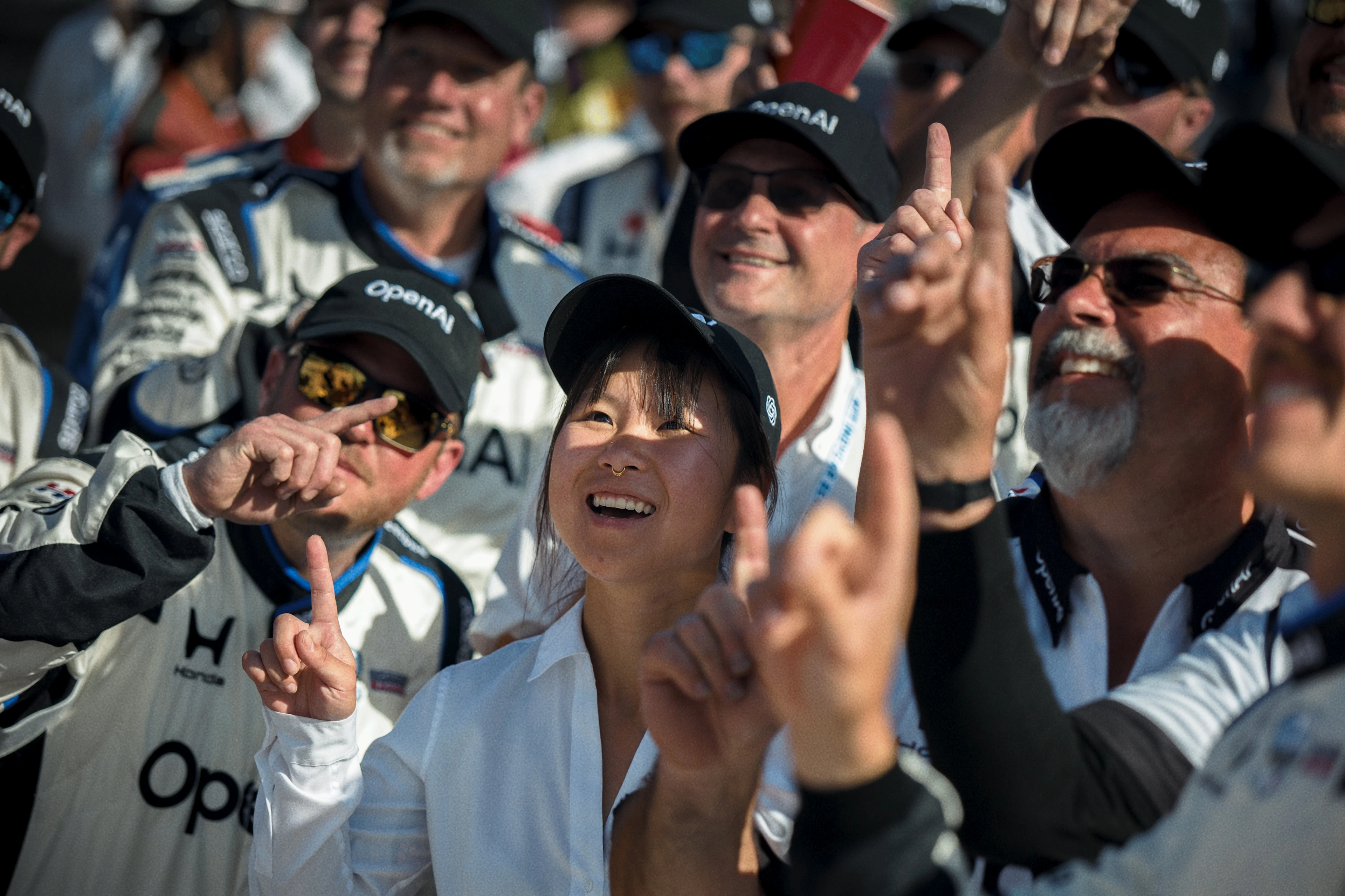 A group of smiling people wearing racing team uniforms and caps gather closely, many raising one finger in celebration or unity.