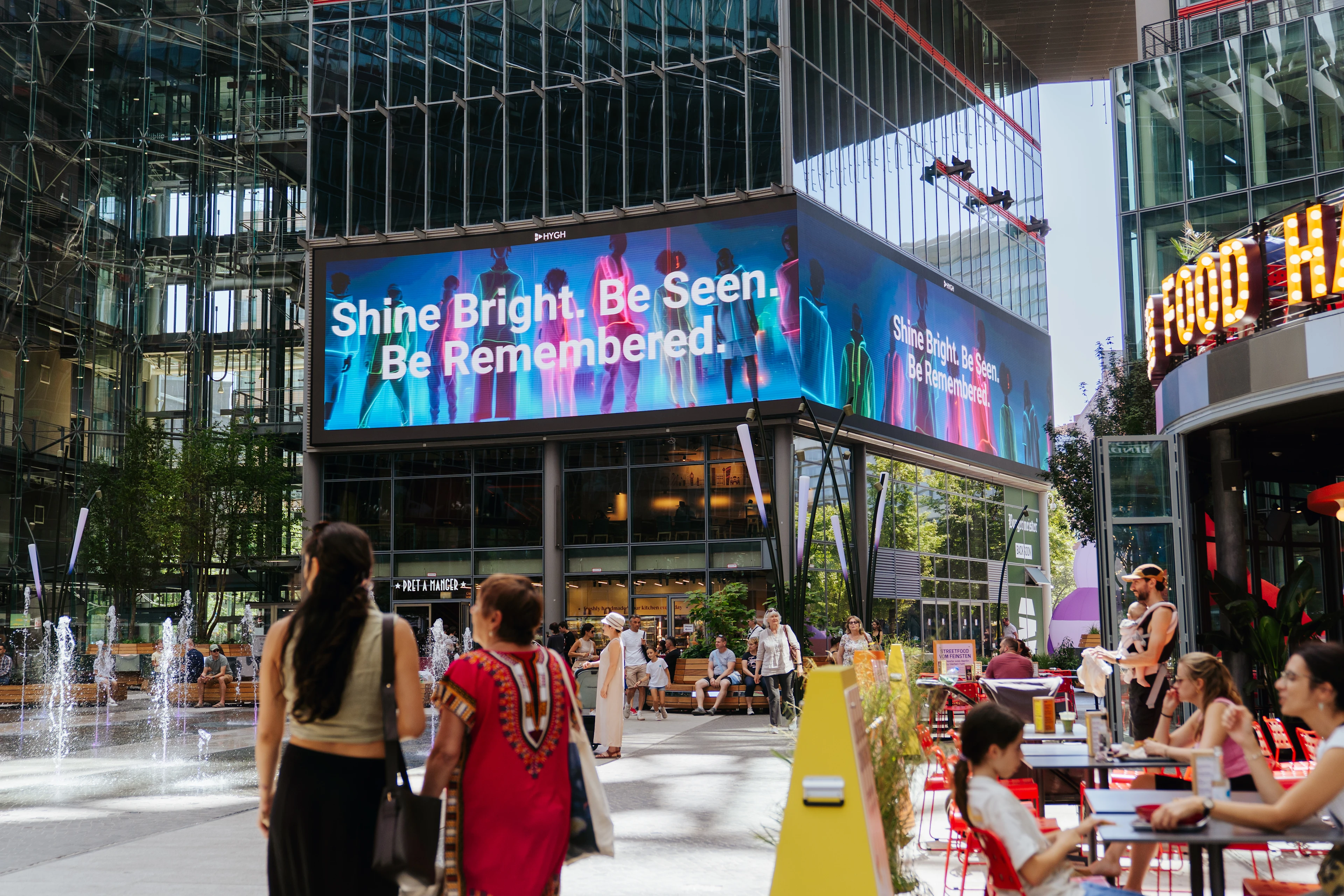 Large HYGH digital billboard at a shopping center displays a vibrant ad reading “Shine Bright. Be Seen. Be Remembered.” The square is filled with people walking, eating, and enjoying a plaza with fountains and cafes.