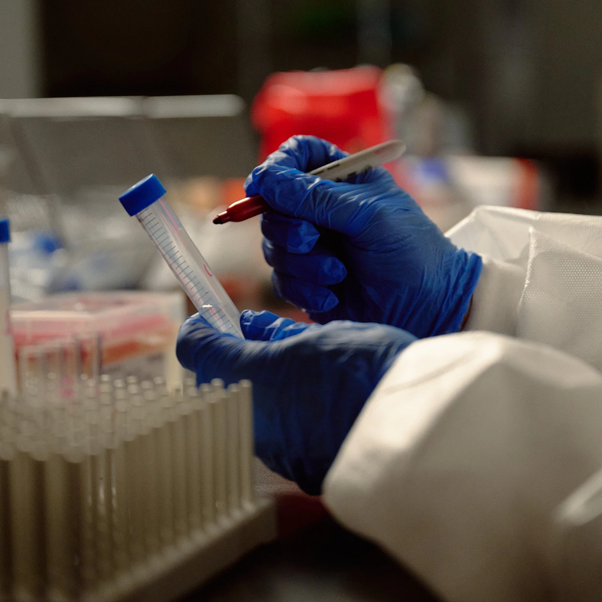 A person wearing blue gloves and a white lab coat labels a plastic test tube with a red marker in a laboratory setting. Several other test tubes are visible in a rack in the foreground, indicating an active scientific or medical testing environment.