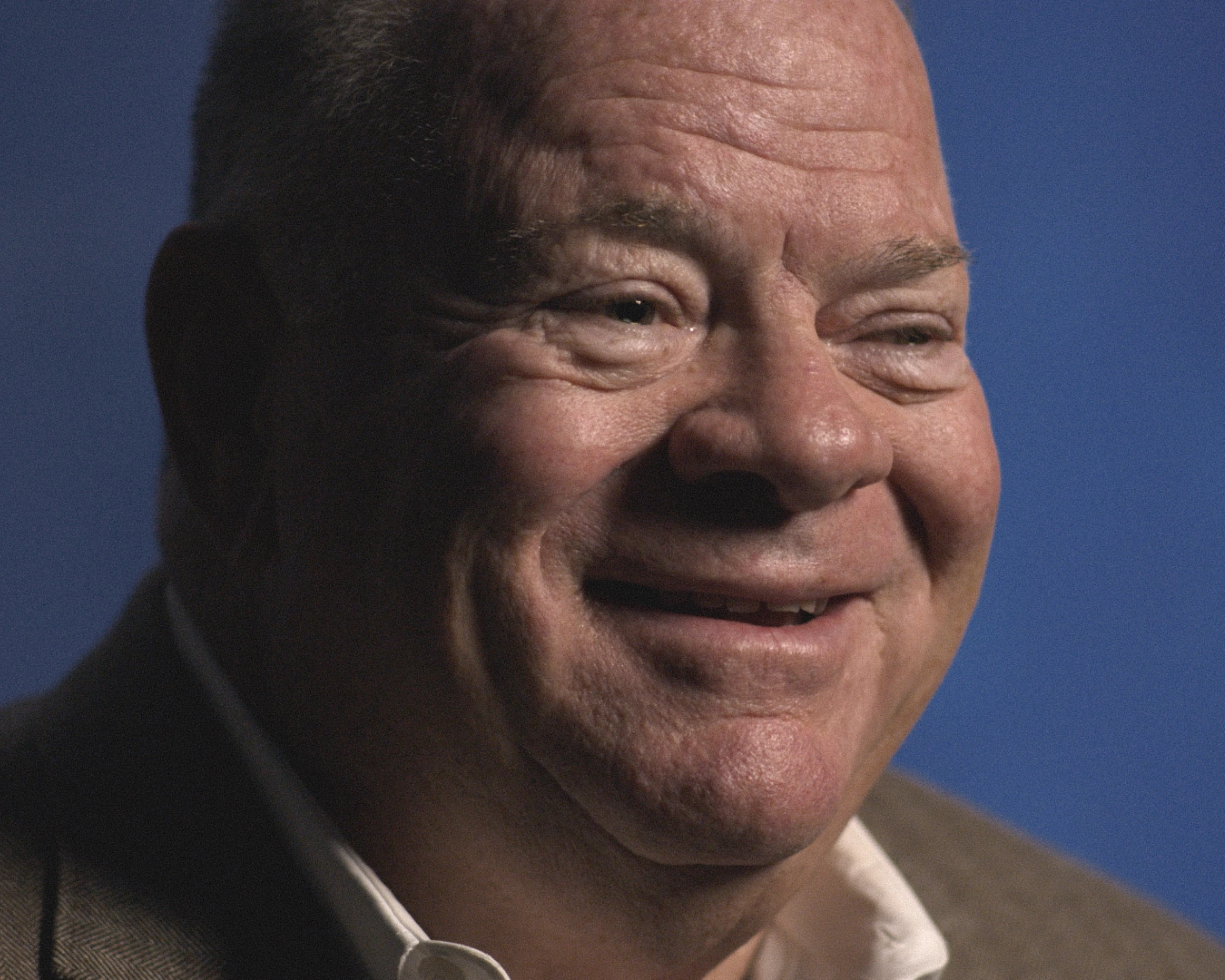 Close-up of an older man with light skin and short hair smiling gently against a plain blue background. He is wearing a brown jacket and white shirt.
