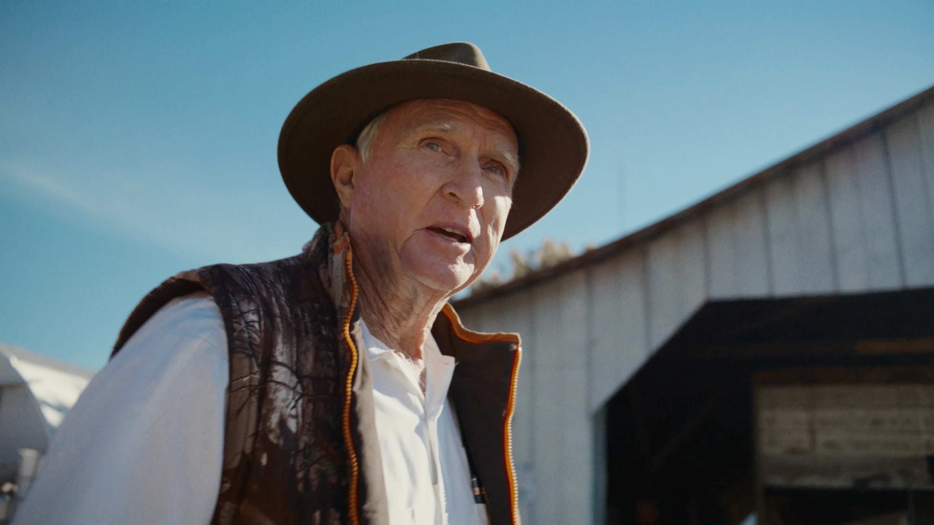 An older man wearing a wide-brimmed hat, white shirt, and brown vest stands outdoors near a farm building under a bright blue sky.