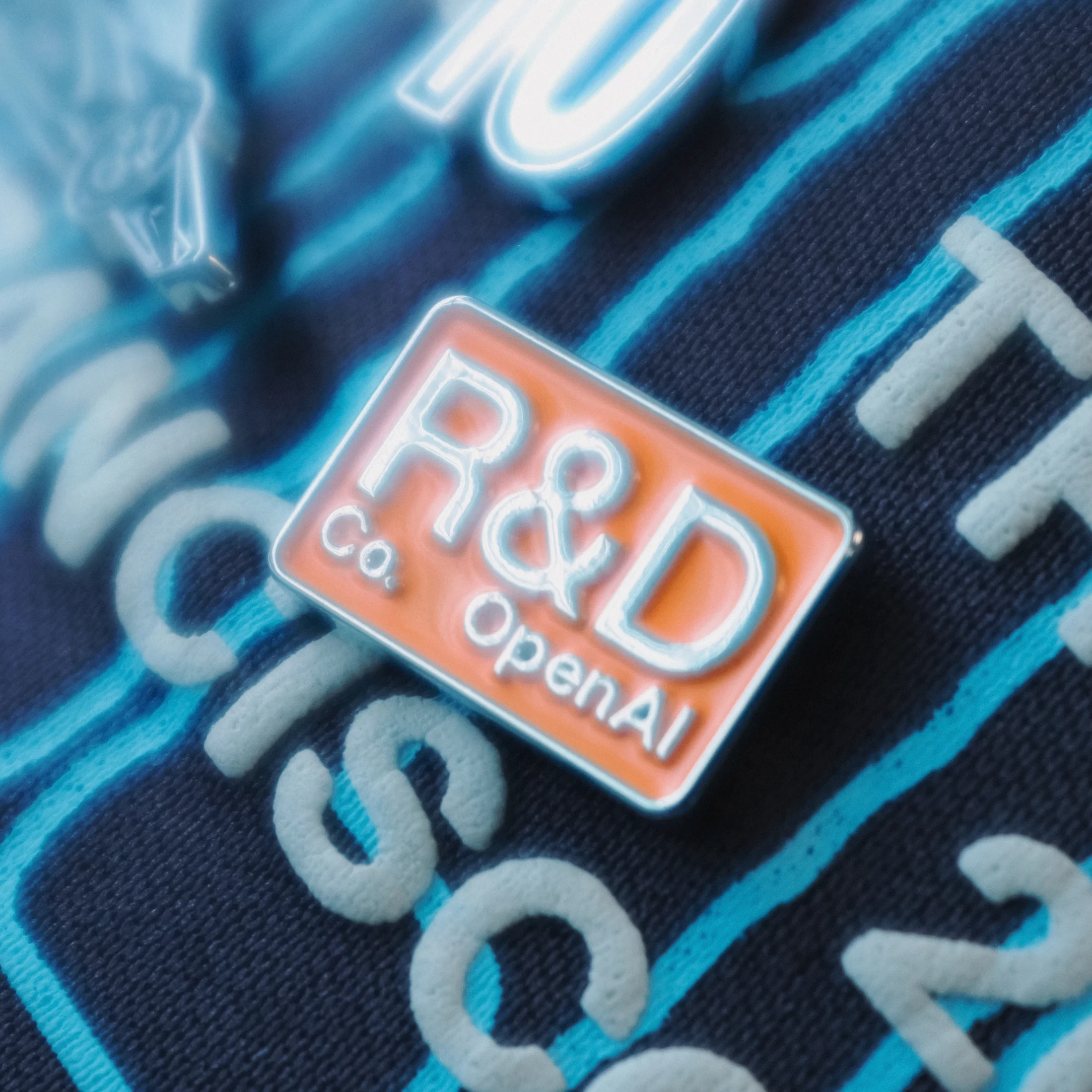An orange R&D Co. OpenAI pin on blue racing merchandise.