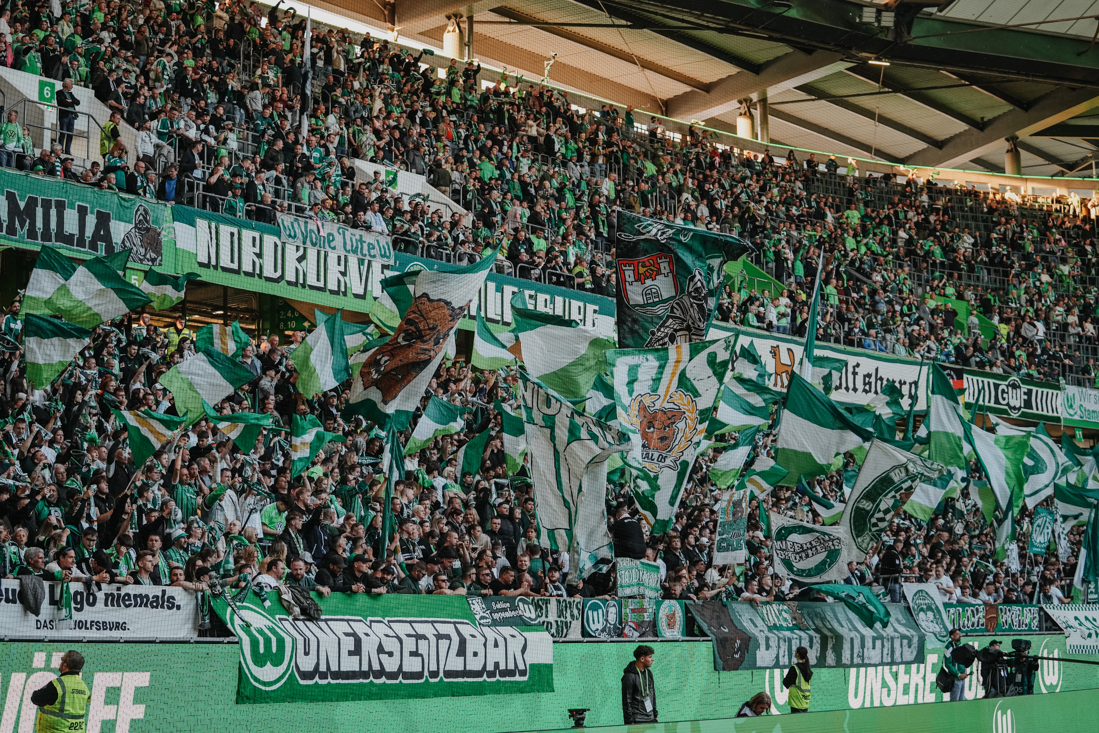Packed stadium stand of VfL Wolfsburg supporters waving green-and-white flags, with large “Nordkurve” and “Unersetzbar” banners stretched across the terraces.