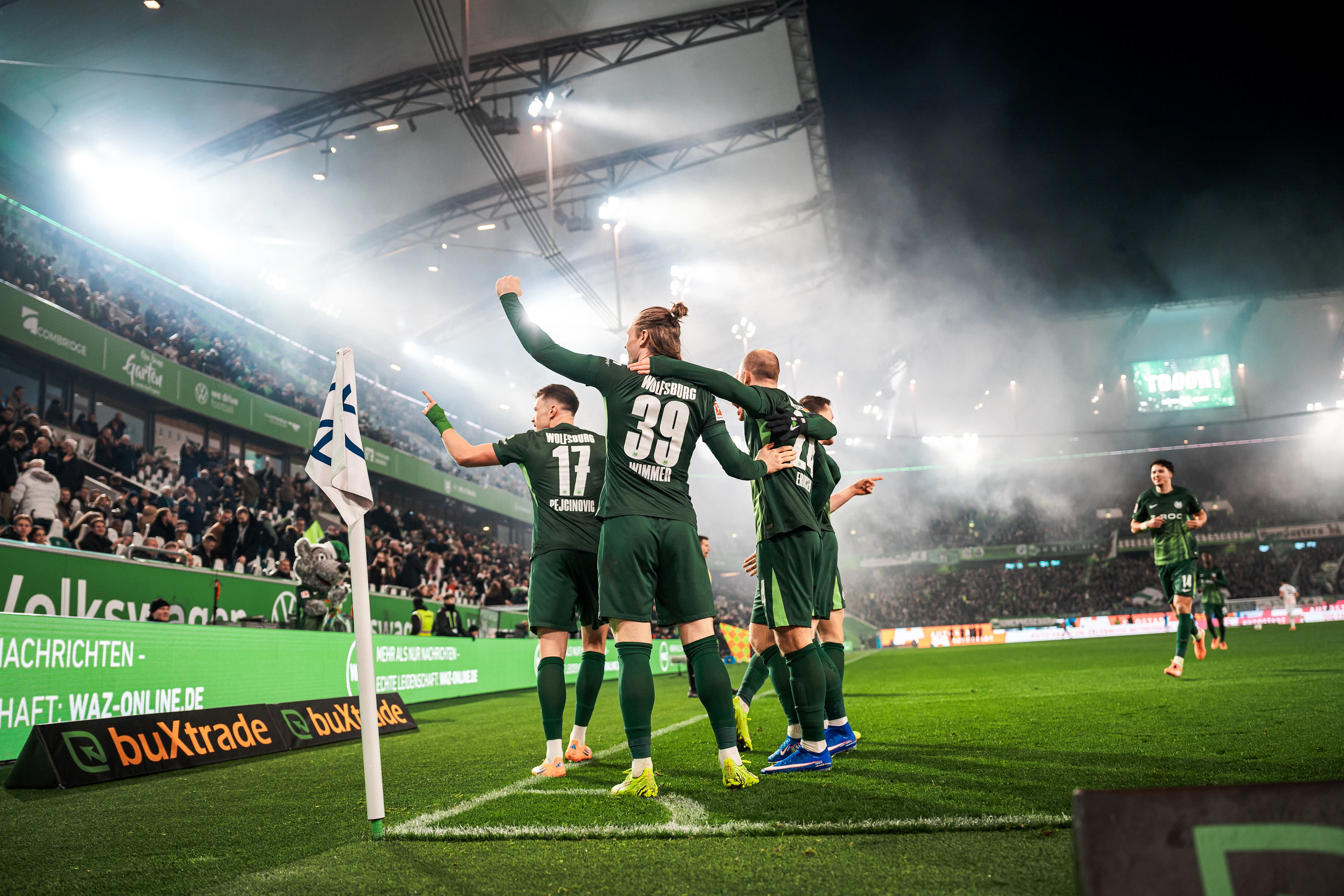 VfL Wolfsburg players celebrate together near the corner flag after scoring, with fans cheering in the stands behind them.