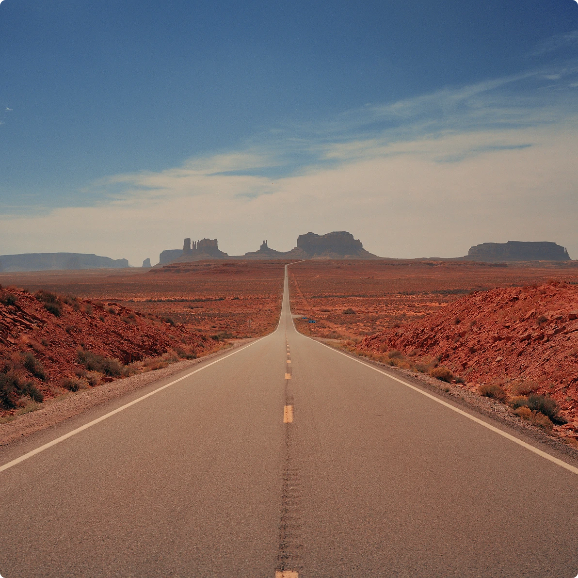 Two-lane highway stretching through a red desert landscape toward distant rock mesas under a wide blue sky.
