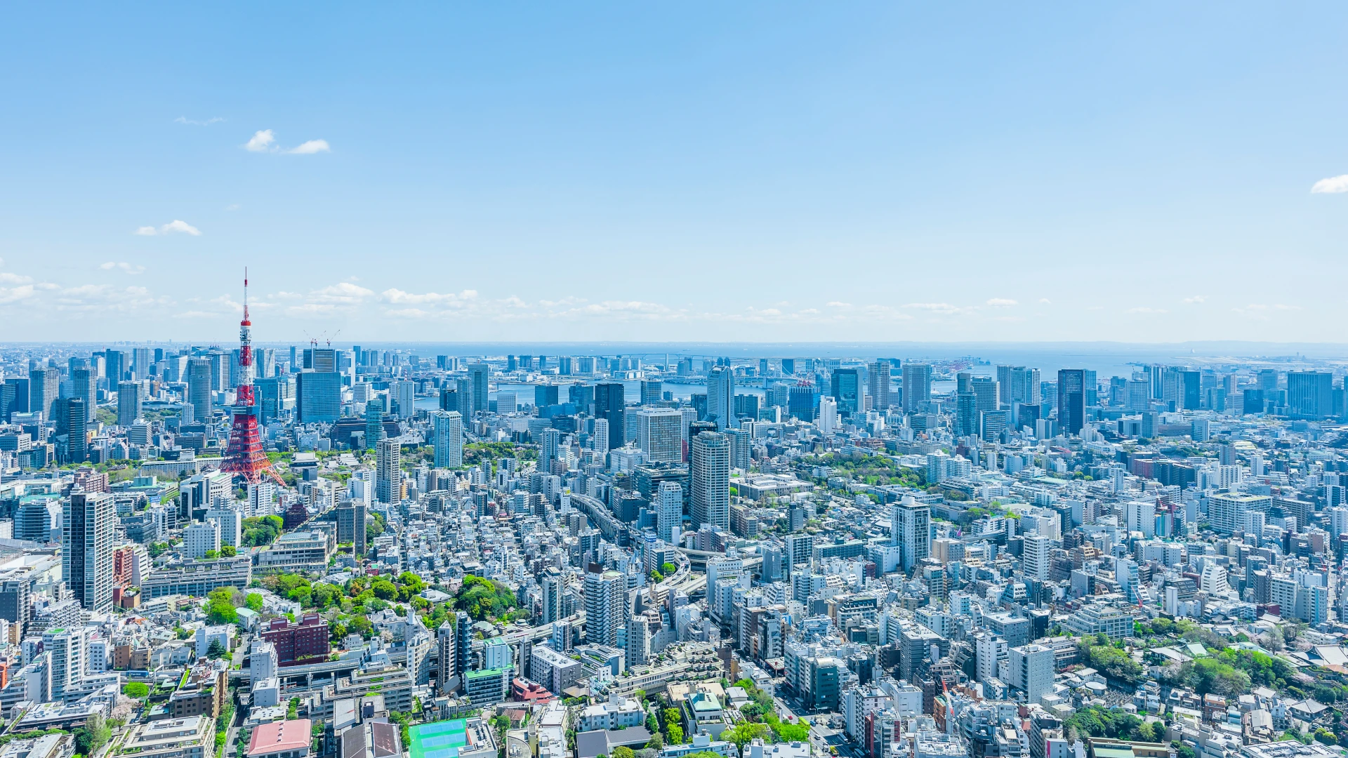 Panoramic daytime view of Tokyo’s skyline, with the red-and-white Tokyo Tower rising above dense city blocks and high-rises under a clear blue sky.
