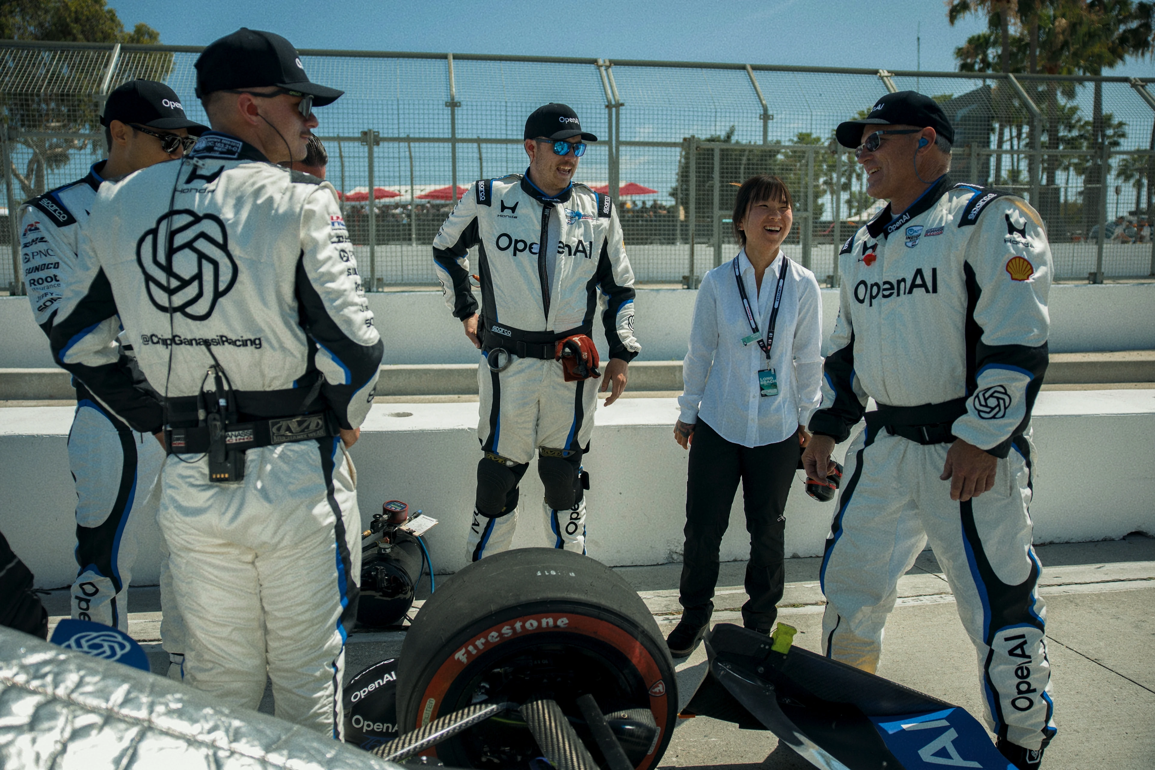A group of five people, four men in white racing suits with OpenAI logos and one woman in a white shirt and black pants, stand and chat beside a race car tire on a race track. They appear relaxed and are smiling, with a metal fence and trees in the background.